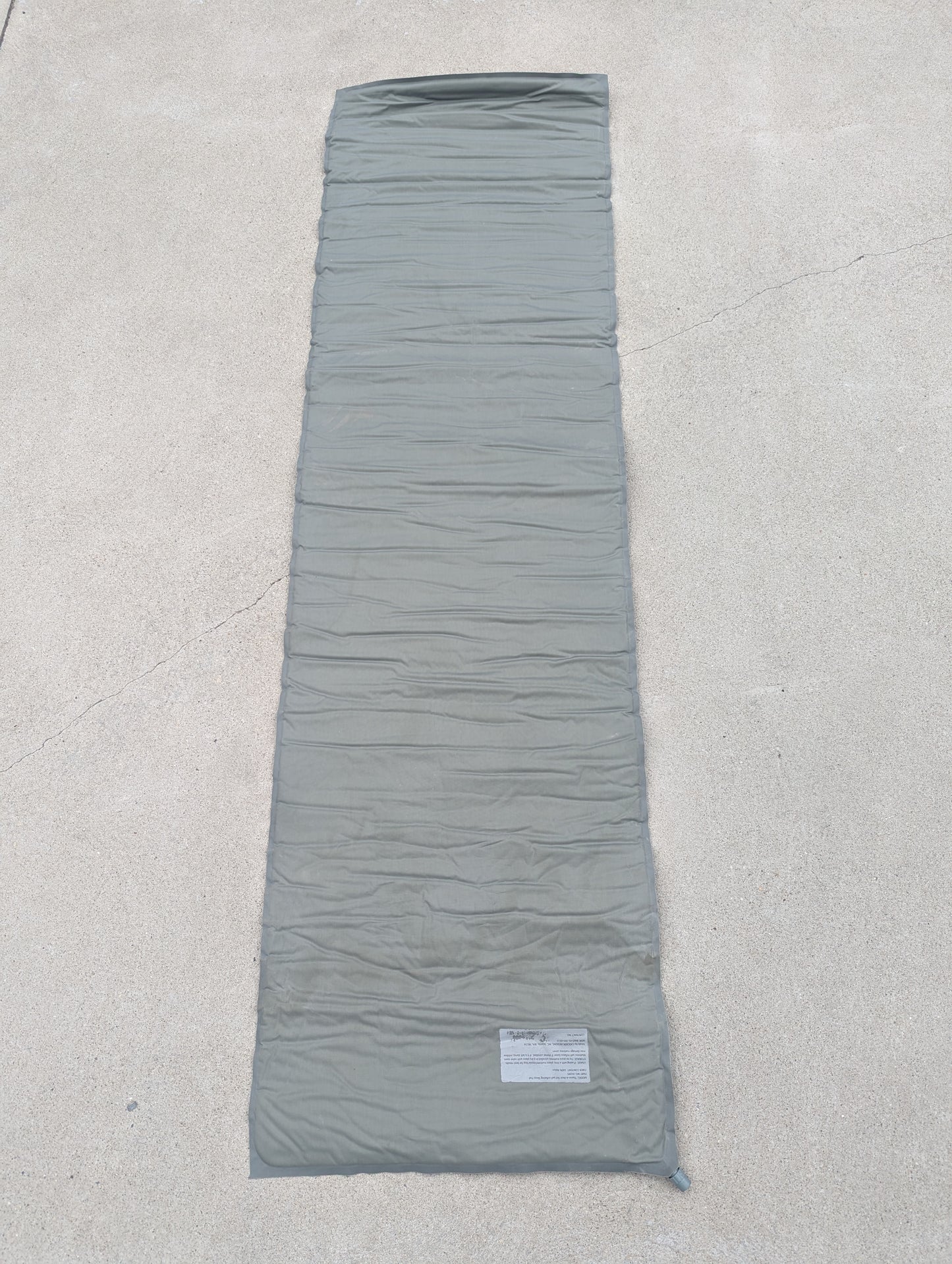 A full-length view of a U.S. Military Surplus self-inflating sleeping mat in foliage green, laid flat on a concrete surface, highlighting the horizontal ribbed pattern and instruction label.