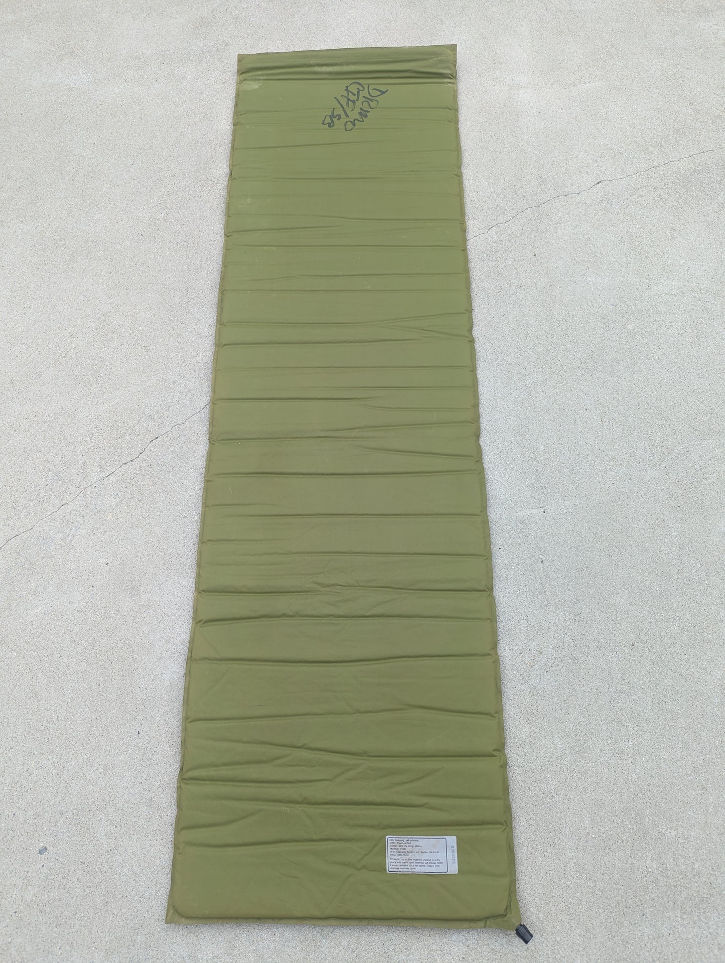 A full-length overhead view of a U.S. Military Surplus self-inflating sleeping mat in foliage green, laid flat on a concrete surface, displaying the horizontal ribbed texture and handwritten surplus markings.