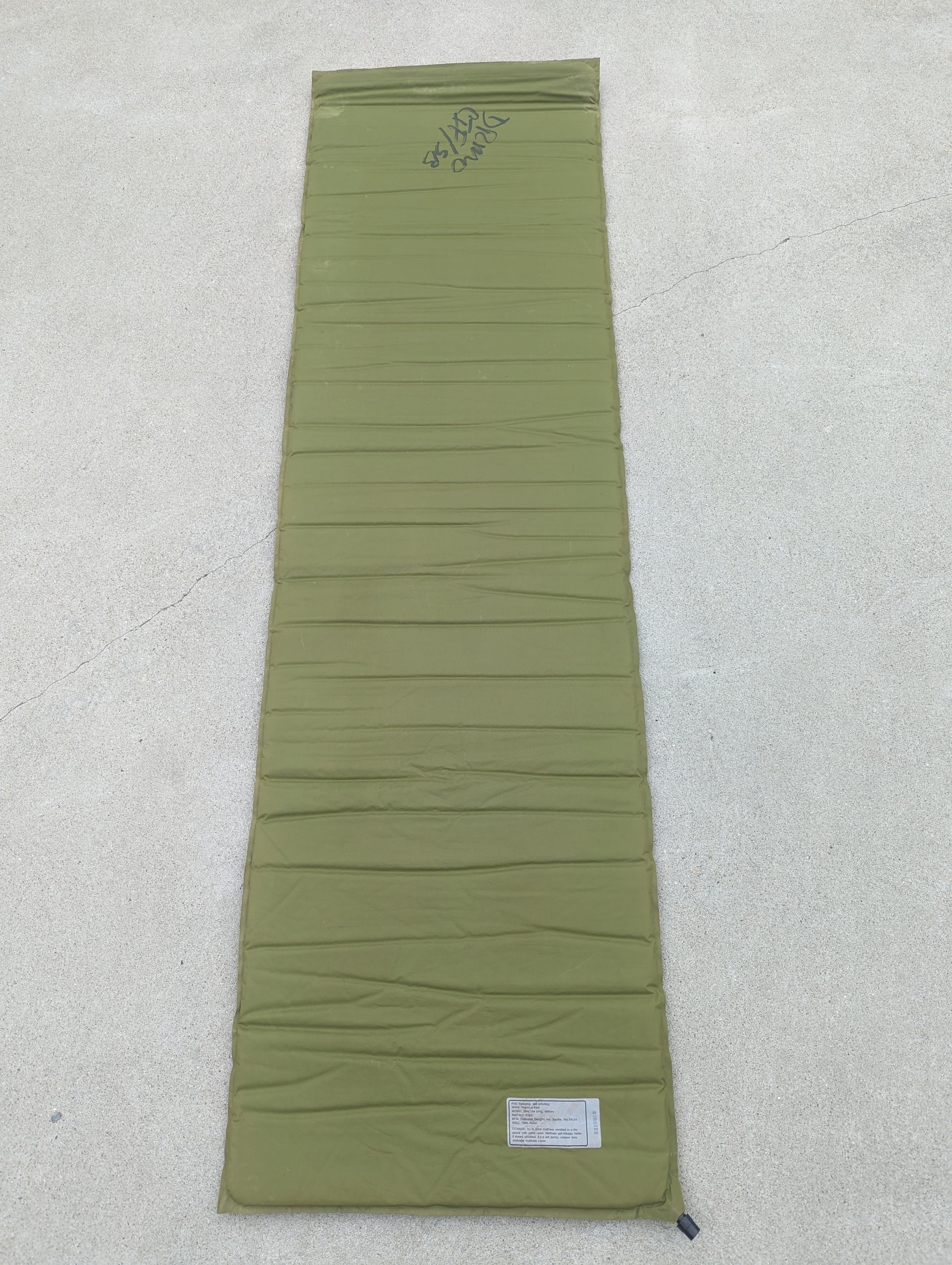 A full-length overhead view of a U.S. Military Surplus self-inflating sleeping mat in foliage green, laid flat on a concrete surface, displaying the horizontal ribbed texture and handwritten surplus markings.