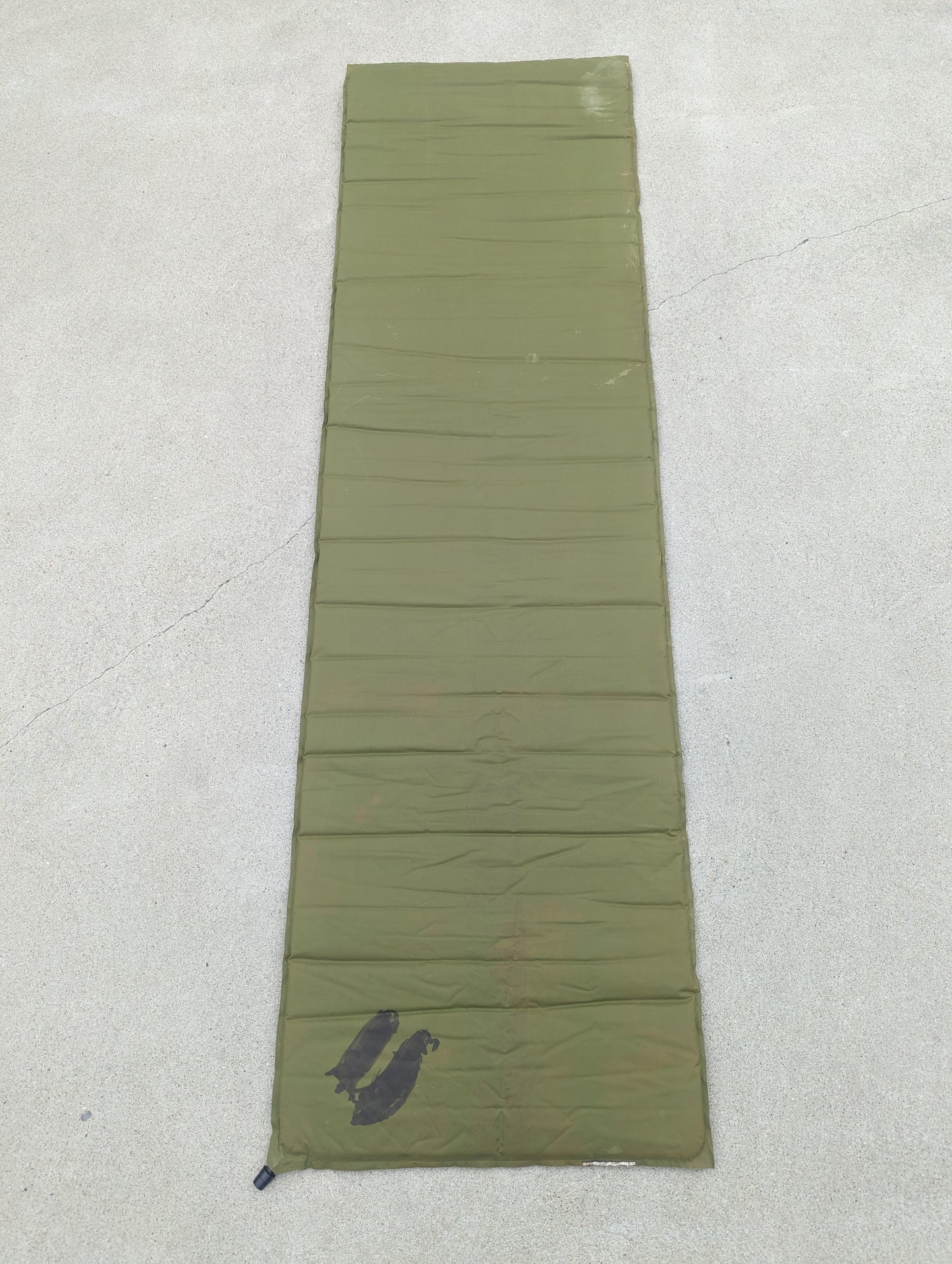 A full-length vertical view of a U.S. Military Surplus self-inflating sleeping mat in olive green, laid flat on a concrete surface, showing a visible dark stain near the bottom.