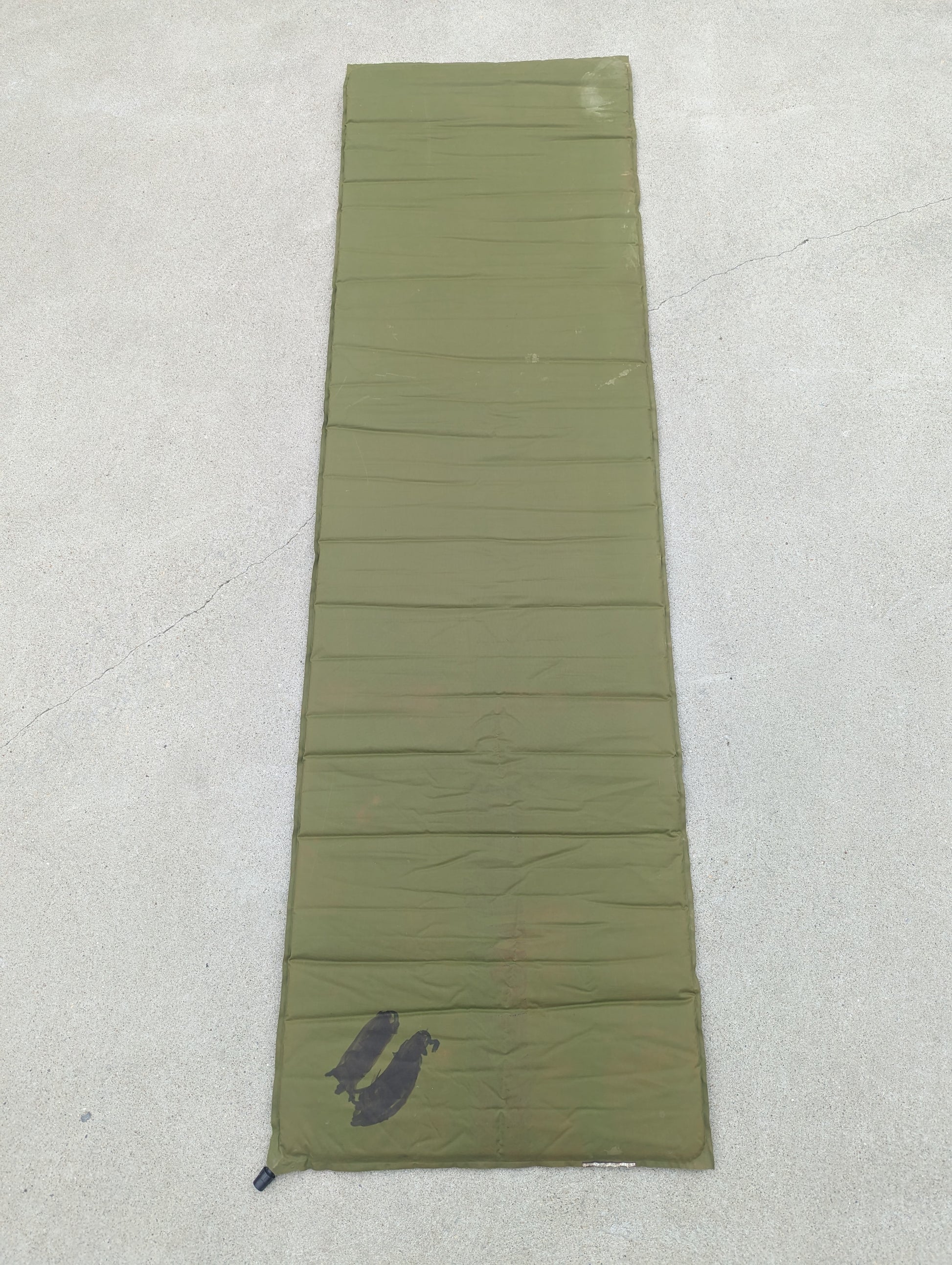 A full-length vertical view of a U.S. Military Surplus self-inflating sleeping mat in olive green, laid flat on a concrete surface, showing a visible dark stain near the bottom.
