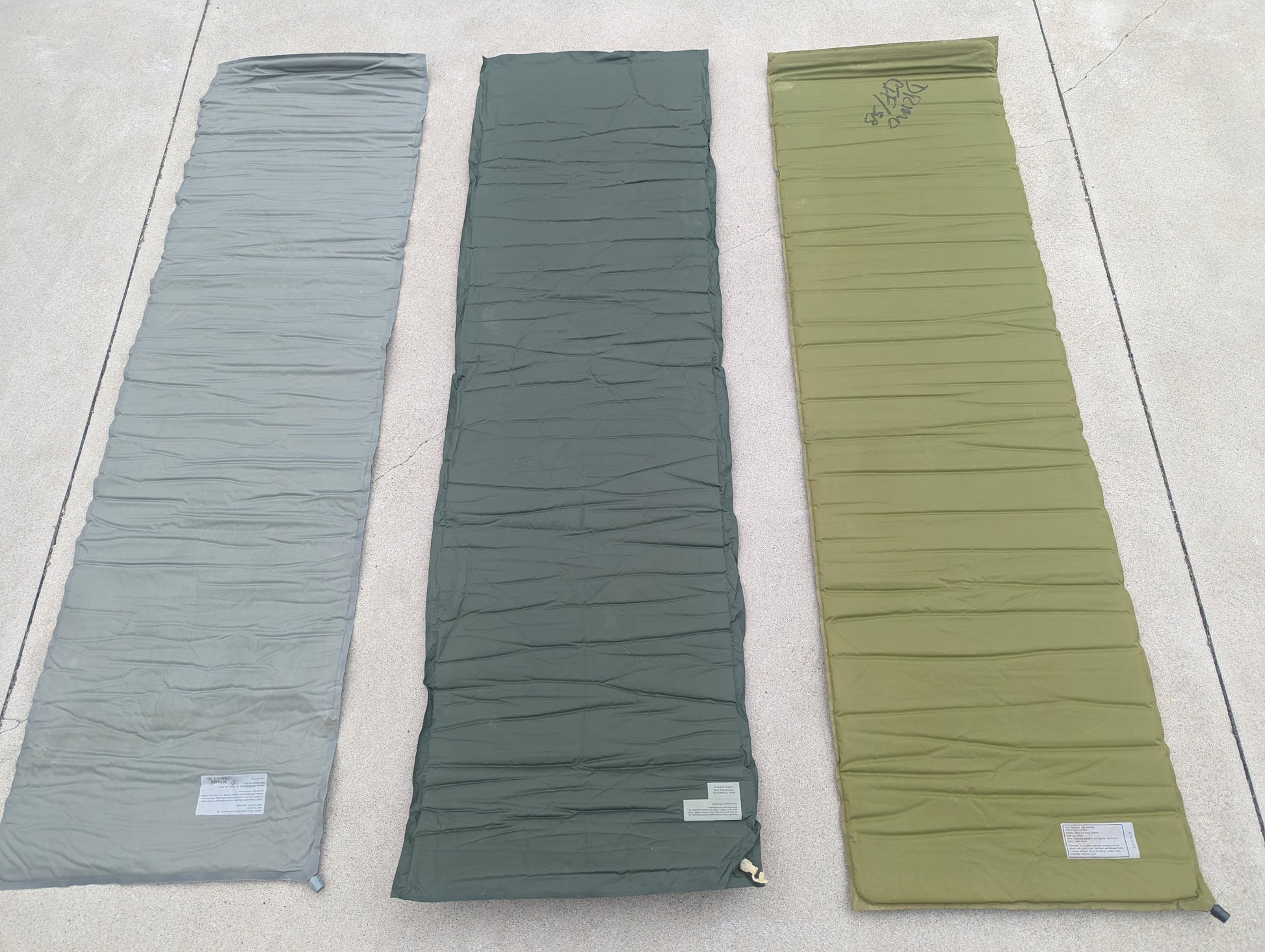 Three U.S. Military surplus self-inflating sleeping mats laid flat side-by-side on a concrete surface, showing color variations in foliage green, dark gray, and olive drab.