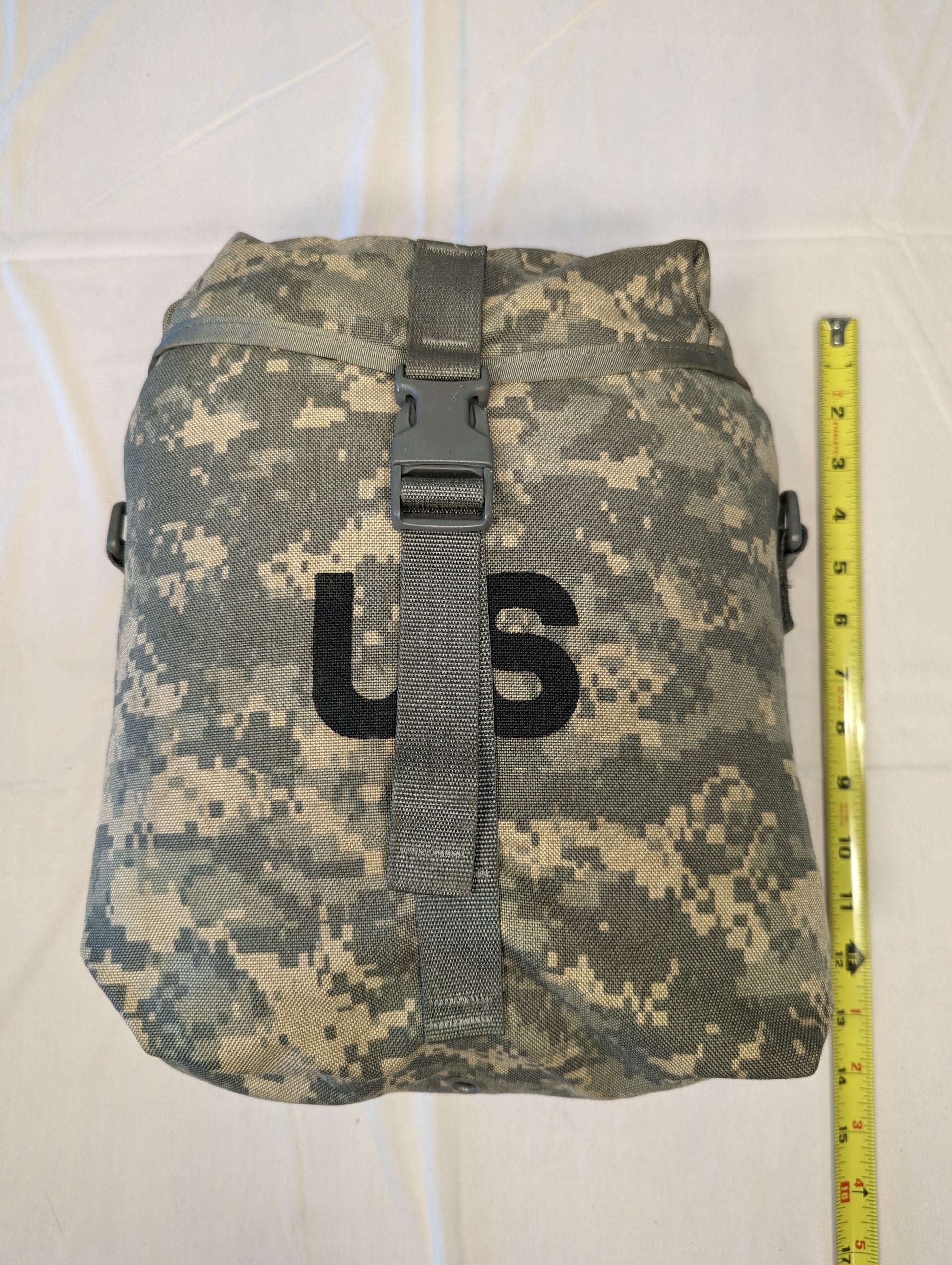 A front view of a U.S. Military Surplus Sustainment Pouch in ACU digital camouflage, standing upright next to a yellow tape measure to demonstrate its height, featuring a large stenciled 'US' mark and a buckle closure.