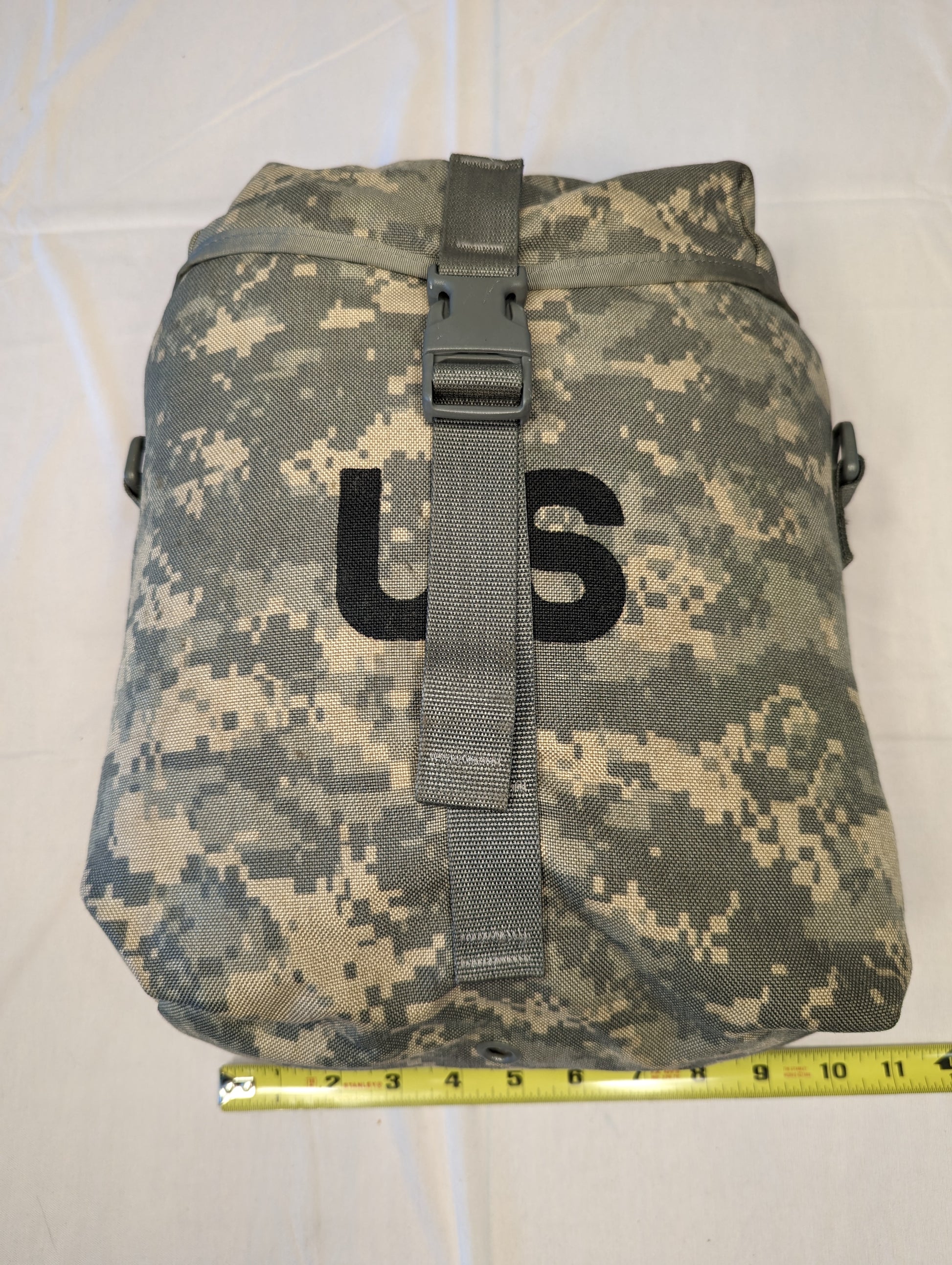 A U.S. Military Surplus Sustainment Pouch in ACU digital camouflage, standing upright, featuring a large black stenciled 'US' mark, a buckle closure, and a yellow tape measure placed at the base to indicate width.