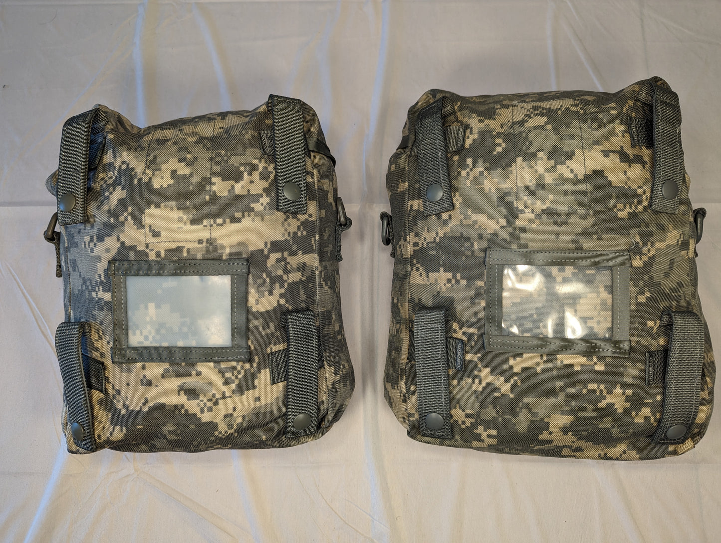 A back view of two U.S. Military Surplus Sustainment Pouches in ACU digital camouflage, laid side-by-side, displaying the vertical MOLLE attachment straps with snap closures and clear vinyl identification windows.