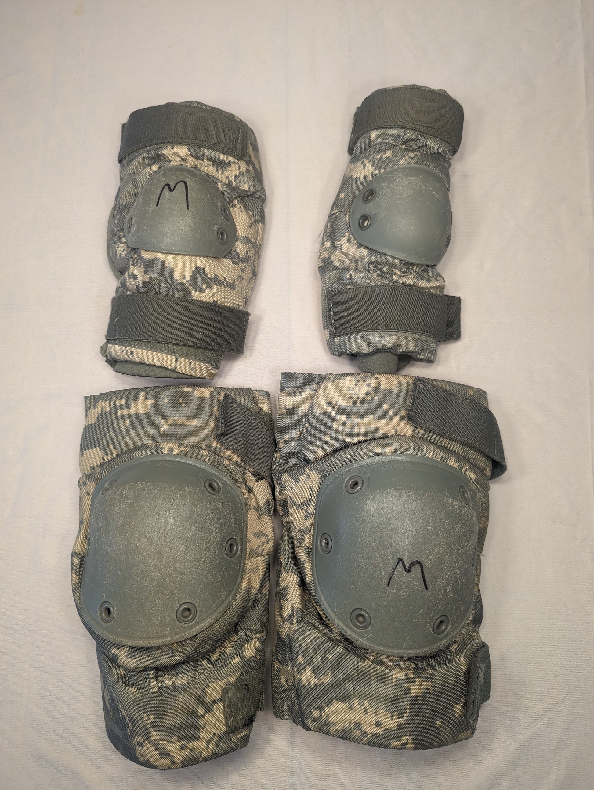 A set of four used U.S. Military tactical knee and elbow pads in ACU digital camouflage, laid out on a white background, featuring dark gray hard plastic shells and black size 'M' markings.