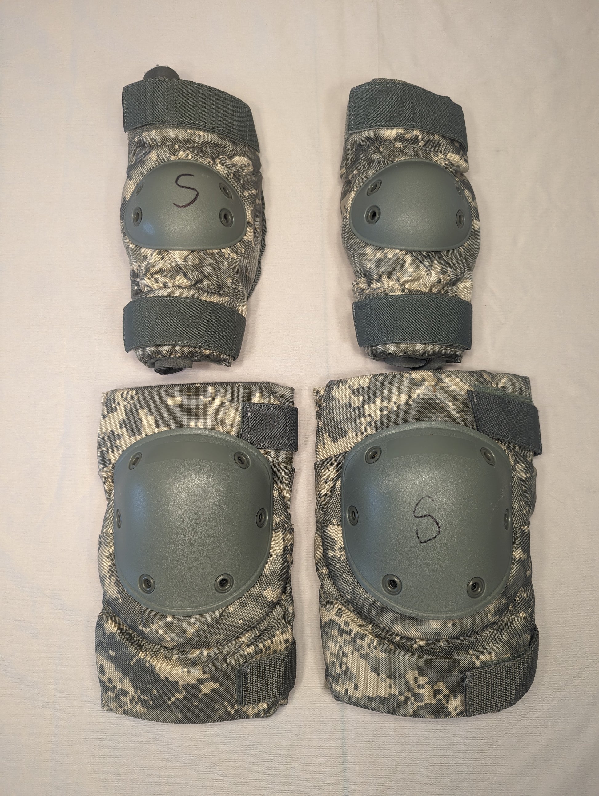A set of four used U.S. Military tactical knee and elbow pads in ACU digital camouflage, laid out on a white background, featuring gray hard plastic shells and black size 'S' markings.