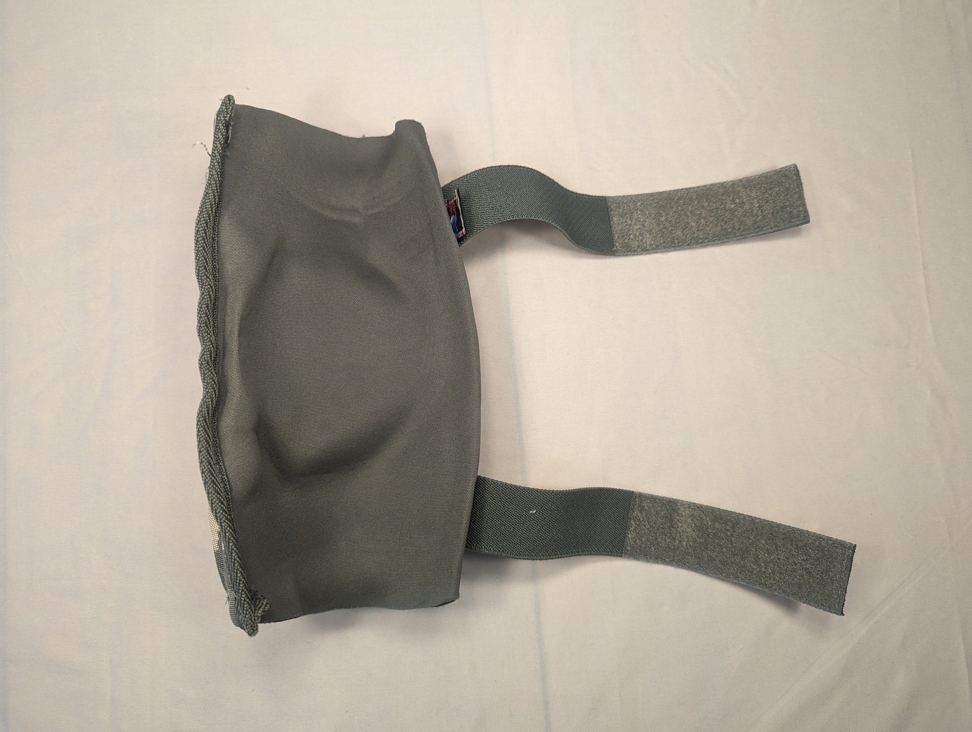 A single used U.S. Military tactical pad in dark gray/foliage green, laid flat, showing the hinged, contoured shape, the attached U.S. flag tag, and the two adjustable hook-and-loop straps.