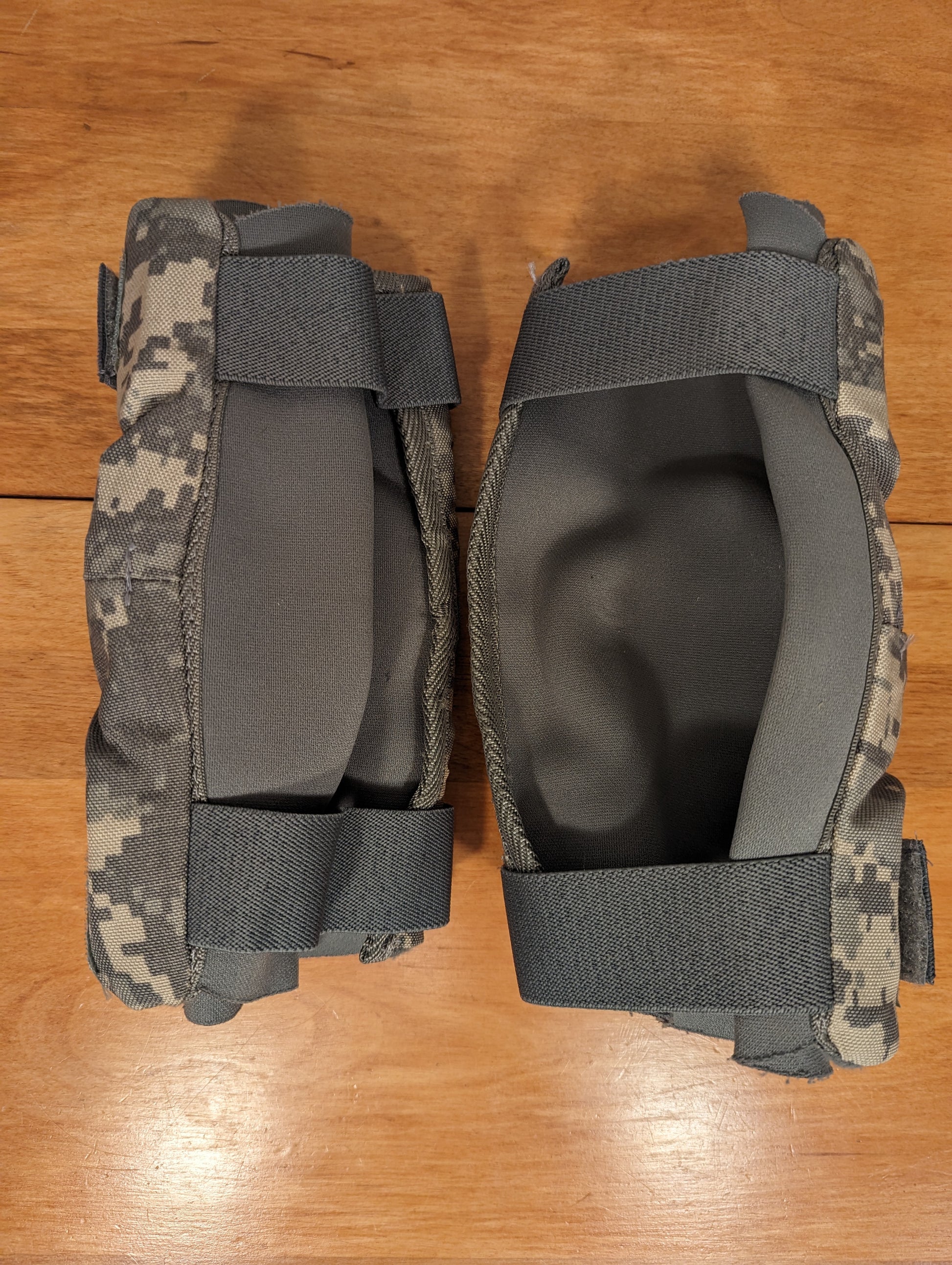 A pair of U.S. Military Surplus Tactical Elbow Pads in ACU digital camouflage, laid on a wooden surface, showing the gray padded inner lining, thick elastic straps, and hinged nylon construction.