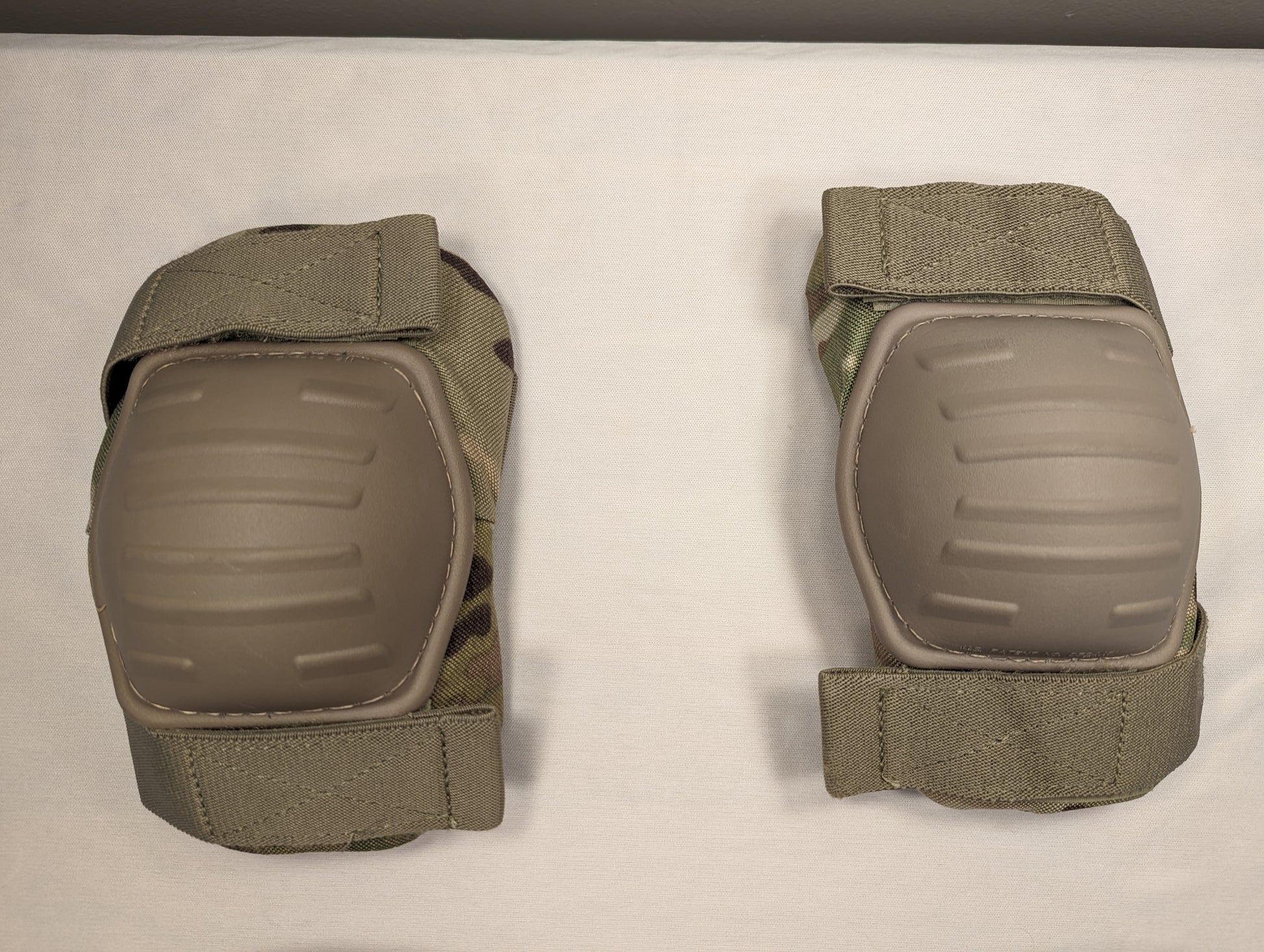 A pair of U.S. Military tactical elbow pads in OCP/Multicam camouflage, laid flat on a white background, featuring tan hard plastic protective shells and adjustable hook-and-loop straps.