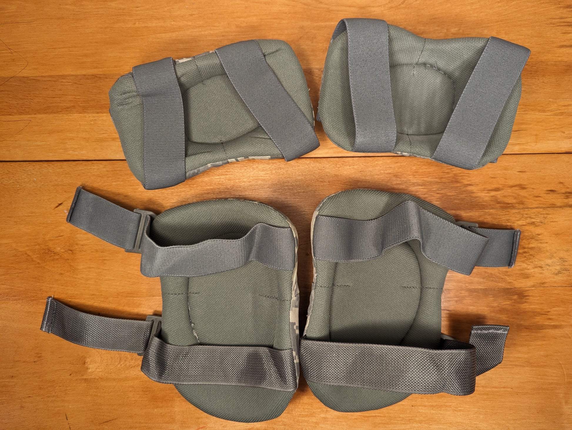A set of four U.S. Military tactical knee and elbow pads in olive green, laid flat on a wooden surface, showing the fabric-covered padded shells and wide adjustable gray webbing straps.