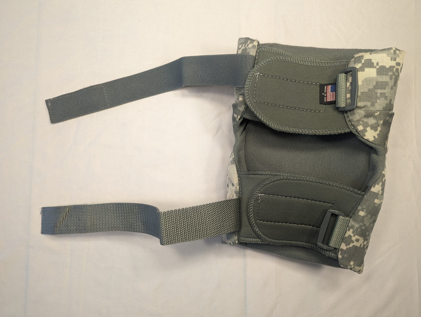 A single used U.S. Military tactical knee or elbow pad in ACU digital camouflage, laid flat, showing the thick padding, wide webbing straps, and the sewn-in U.S. flag tag.