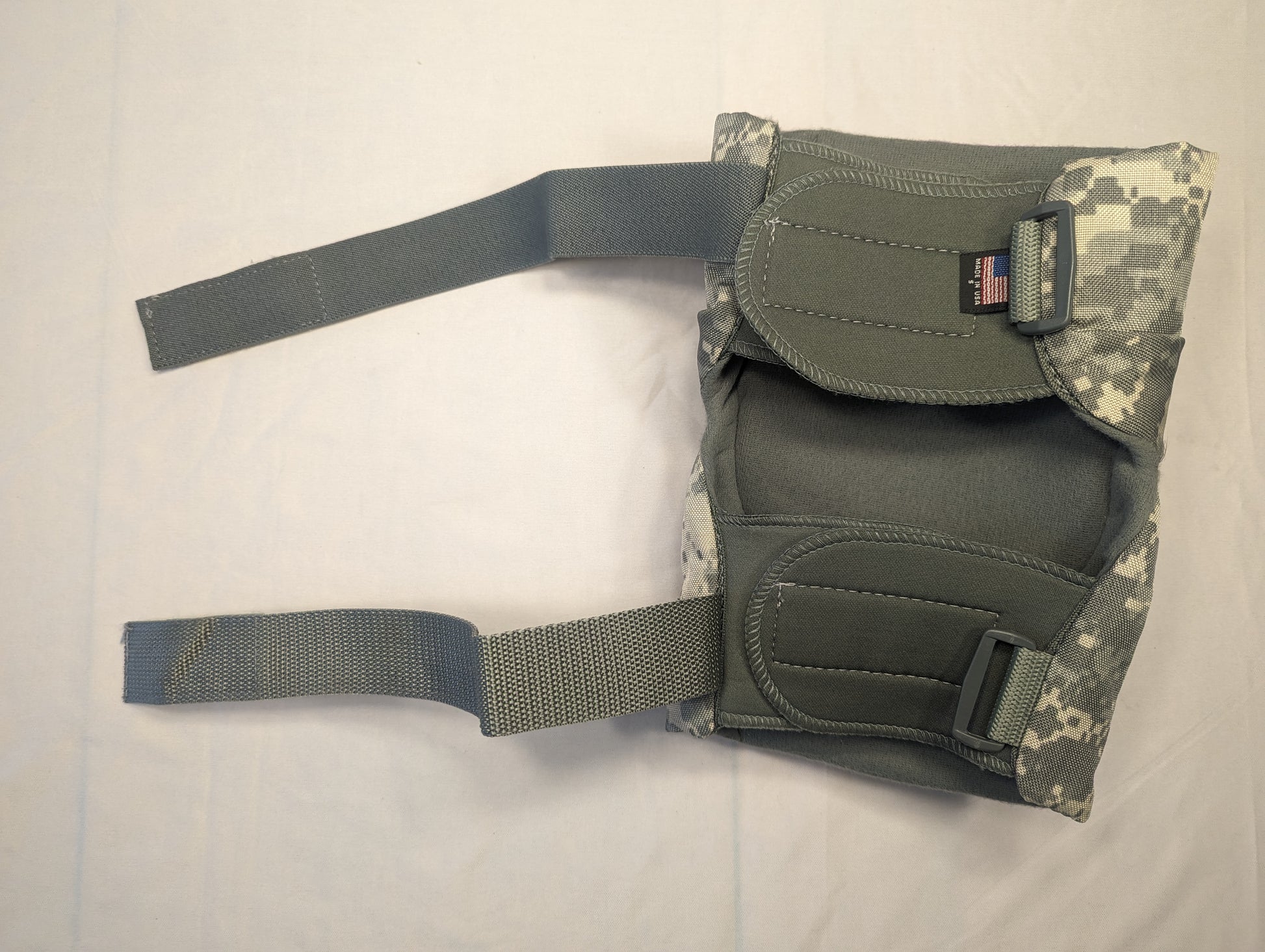 A single used U.S. Military tactical knee or elbow pad in ACU digital camouflage, laid flat, showing the thick padding, wide webbing straps, and the sewn-in U.S. flag tag.