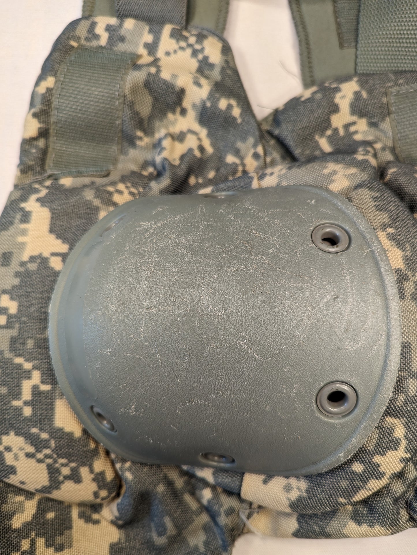 A close-up view of a U.S. Military tactical knee pad in ACU digital camouflage, featuring a foliage green hard plastic protective shell secured with metal grommets.
