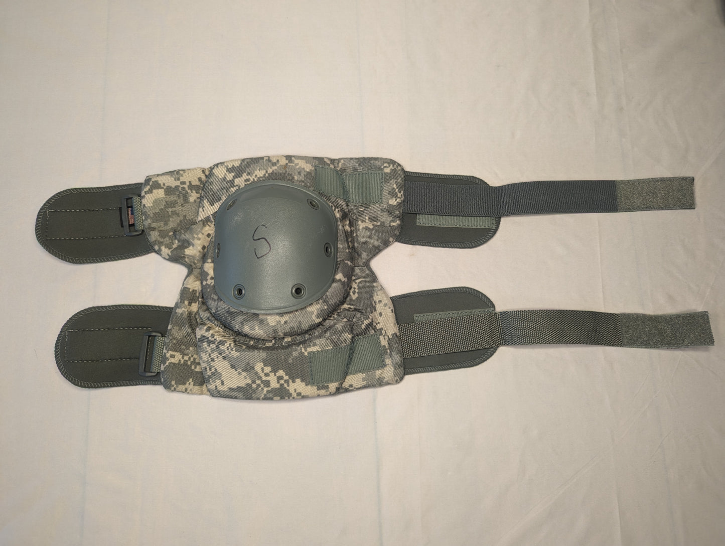 A single U.S. Military tactical knee pad laid flat, featuring an olive green padded fabric backing, a dark gray hard plastic shell marked with an 'S' for size, and ACU digital camouflage straps.