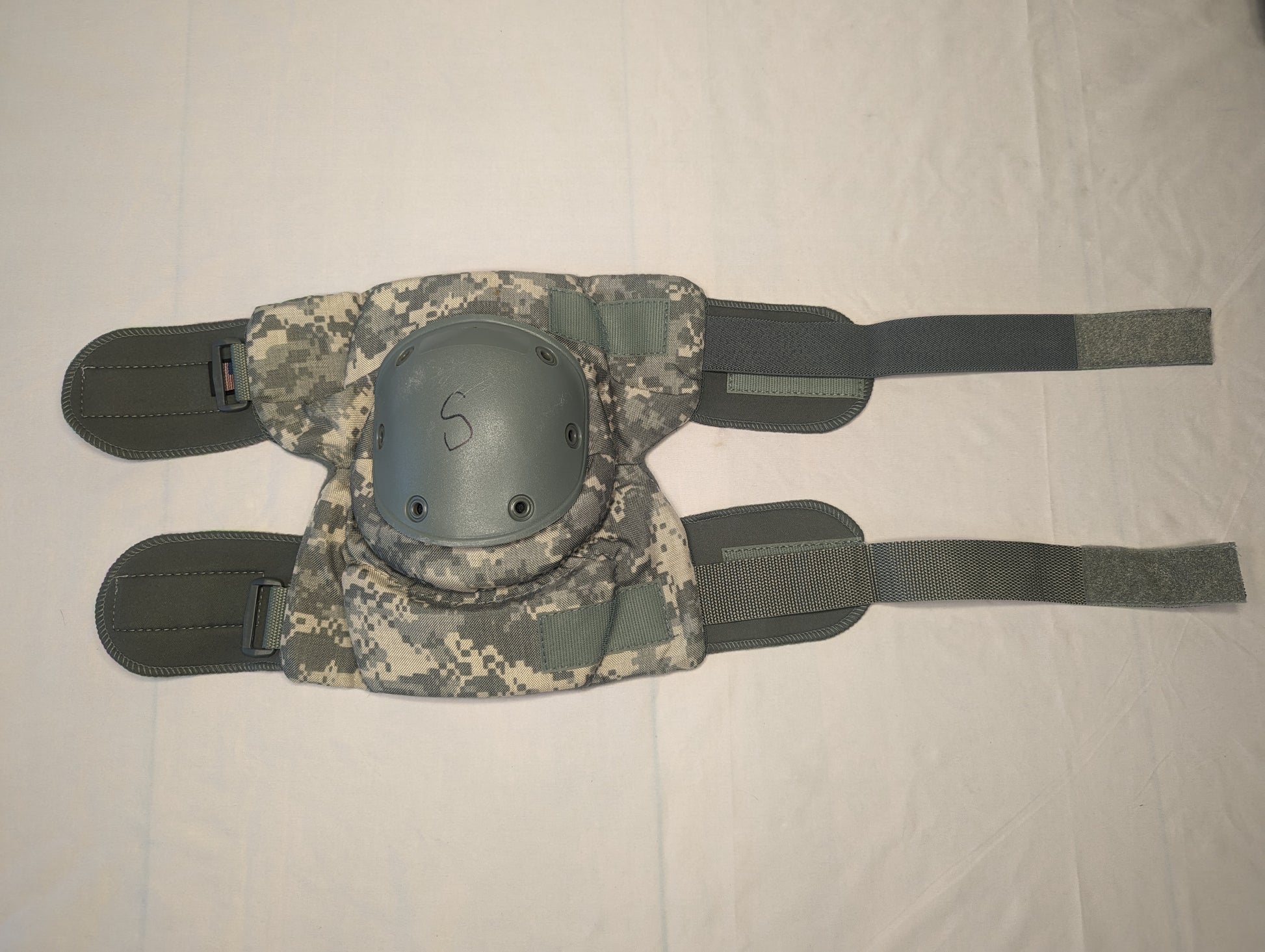 A single U.S. Military tactical knee pad laid flat, featuring an olive green padded fabric backing, a dark gray hard plastic shell marked with an 'S' for size, and ACU digital camouflage straps.