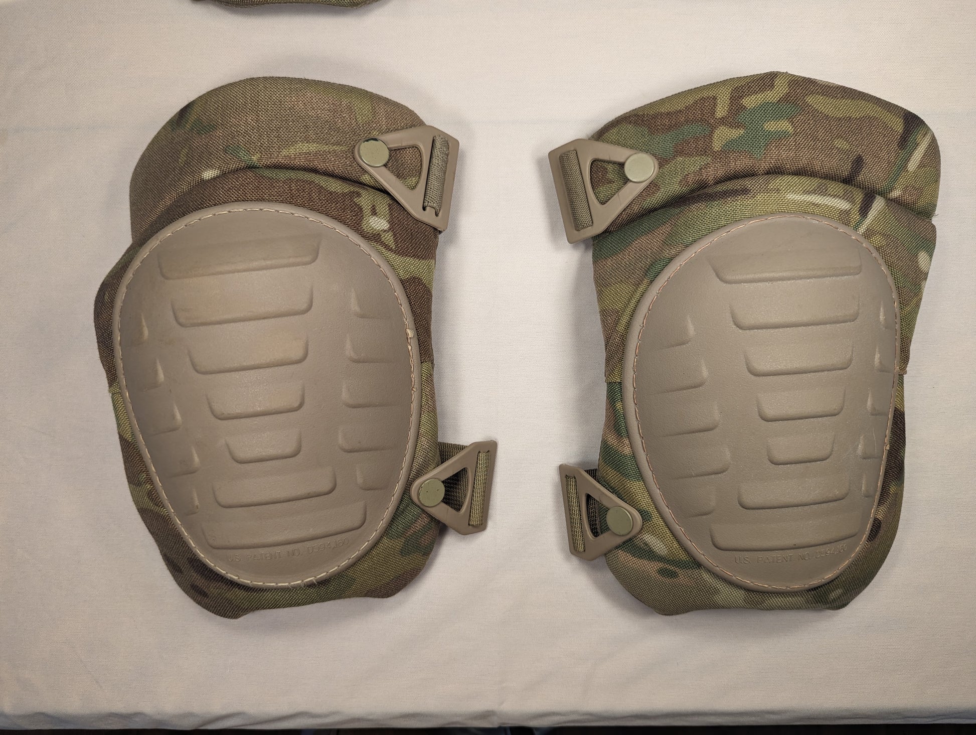 A pair of U.S. Military tactical knee pads in OCP/Multicam camouflage, laid flat side-by-side on a light background, featuring tan hard plastic protective shells and quick-release strap attachments.