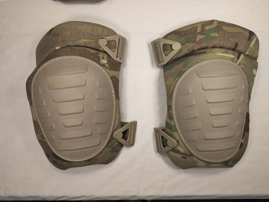 A pair of U.S. Military tactical knee pads in OCP/Multicam camouflage, laid flat side-by-side on a light background, featuring tan hard plastic protective shells and quick-release strap attachments.