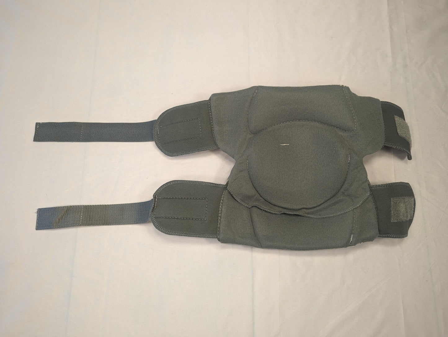 A pair of used tactical knee pads in solid olive green, laid flat on a light surface, showing the heavily padded fabric and the full-length hook-and-loop strap system.