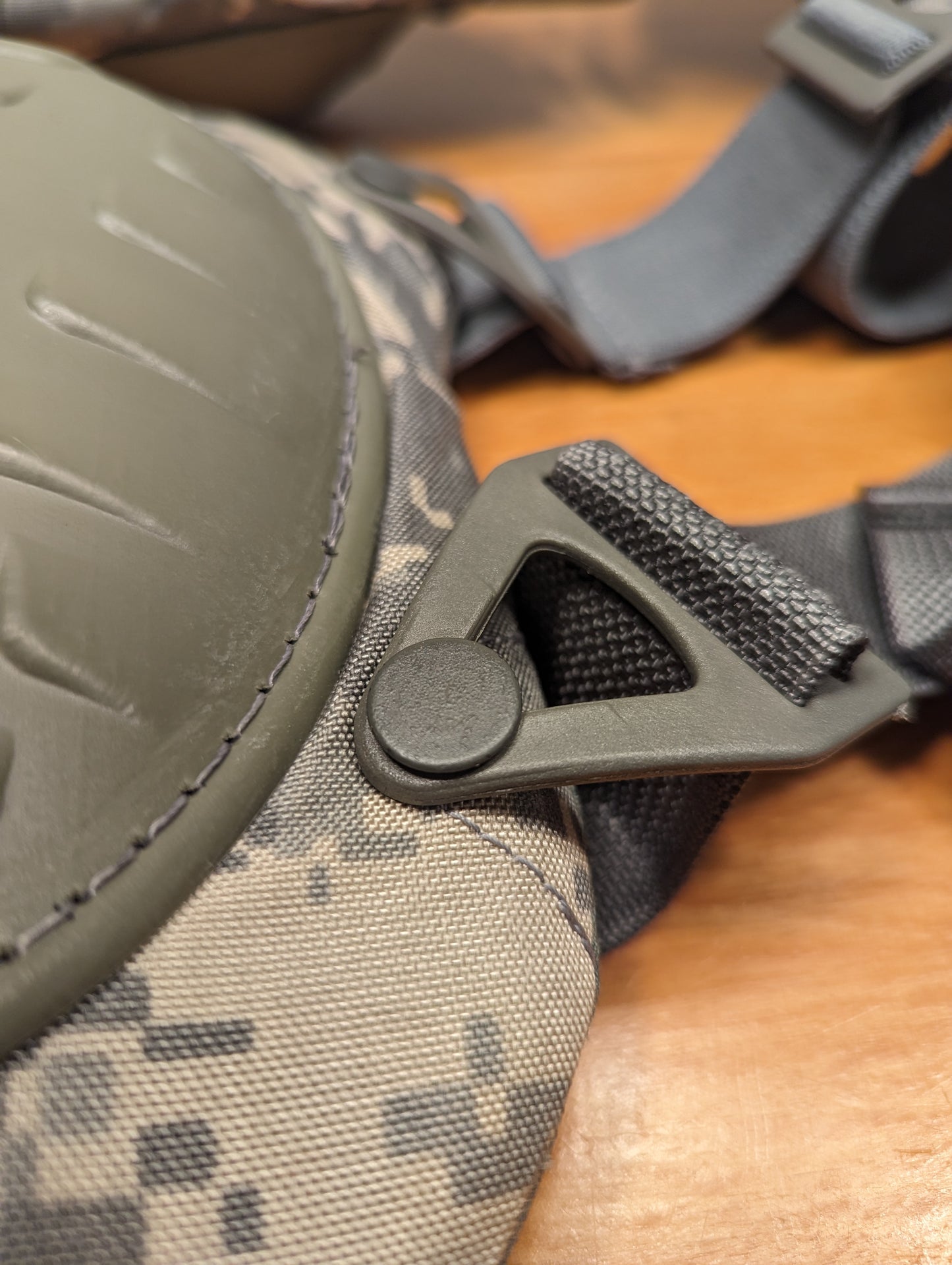 An extreme close-up of the hardware on a U.S. Military tactical knee or elbow pad, highlighting the triangular plastic buckle secured by a rivet onto the ACU digital camouflage fabric and foliage green hard shell.