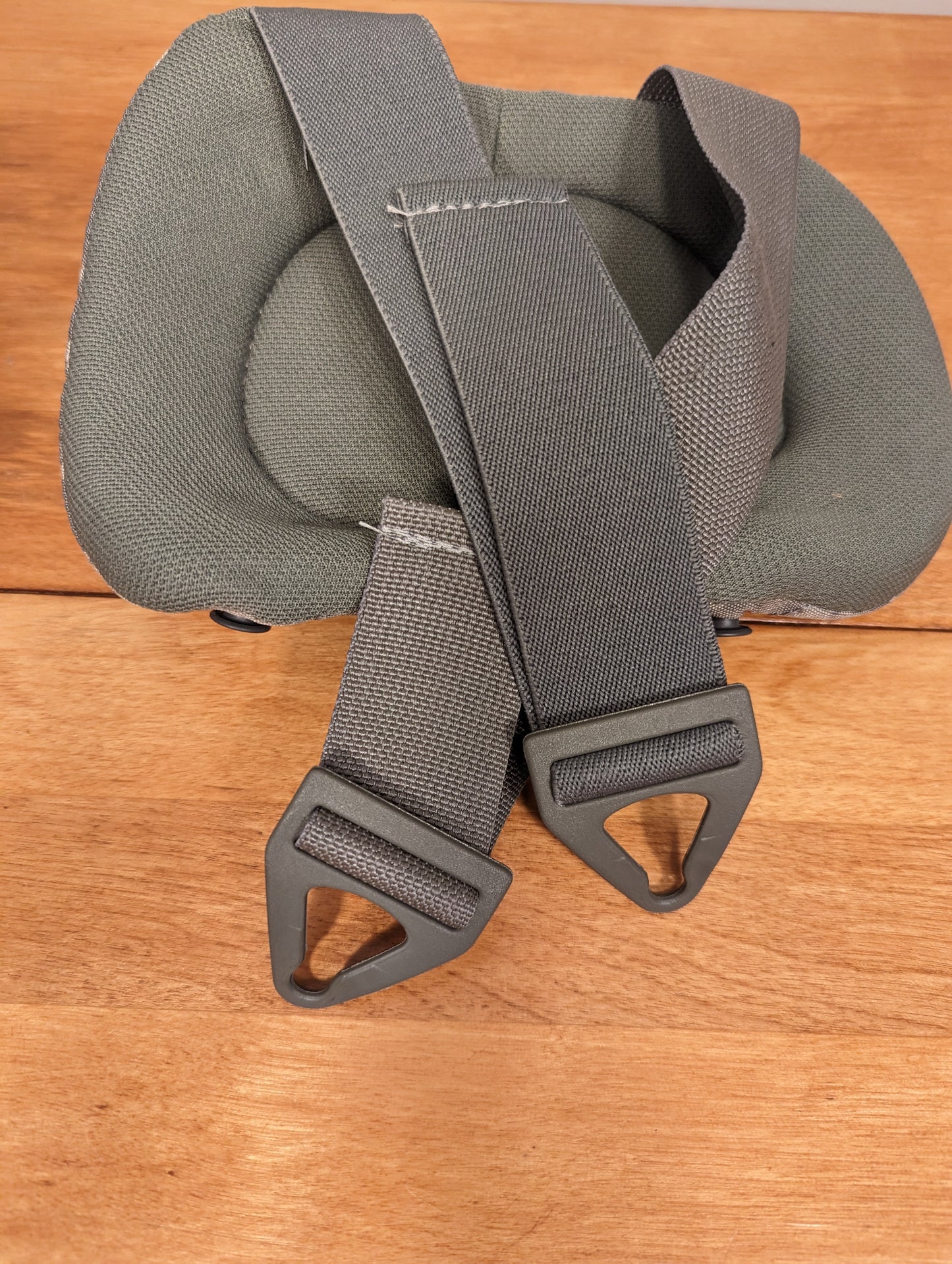 A close-up view of a U.S. Military tactical pad insert in foliage green, laid on a wooden surface, showing the thick padding, gray webbing straps, and two triangular plastic D-ring adjusters.