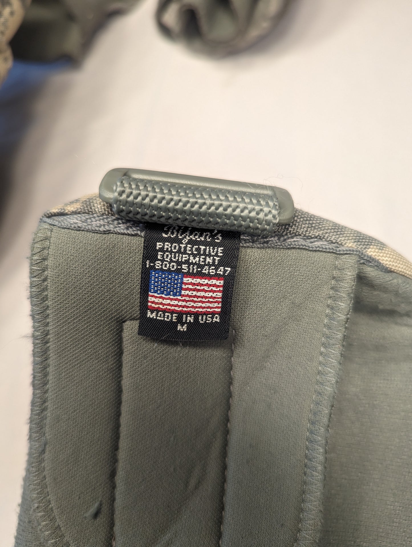 An extreme close-up of the U.S. flag tag and a plastic adjustment buckle sewn onto the foliage green strap of a tactical pad, clearly displaying 'MADE IN U.S.A.' text.