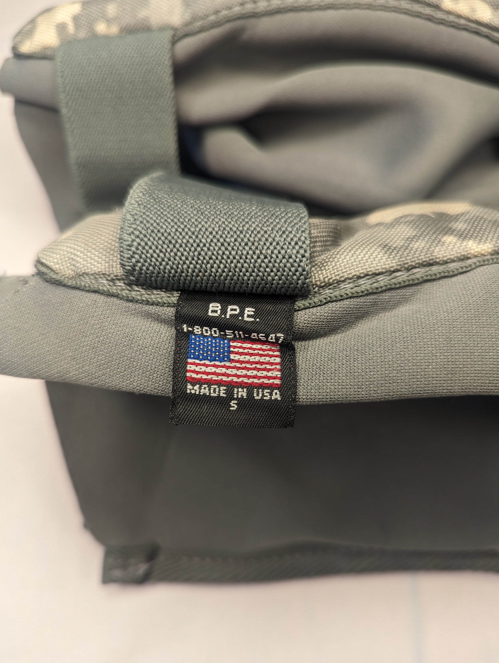 A close-up photograph of the nylon adjustment strap of a tactical pad, highlighting the sewn-in U.S. flag tag with 'MADE IN U.S.A.' text and contractor markings 'BPE'.