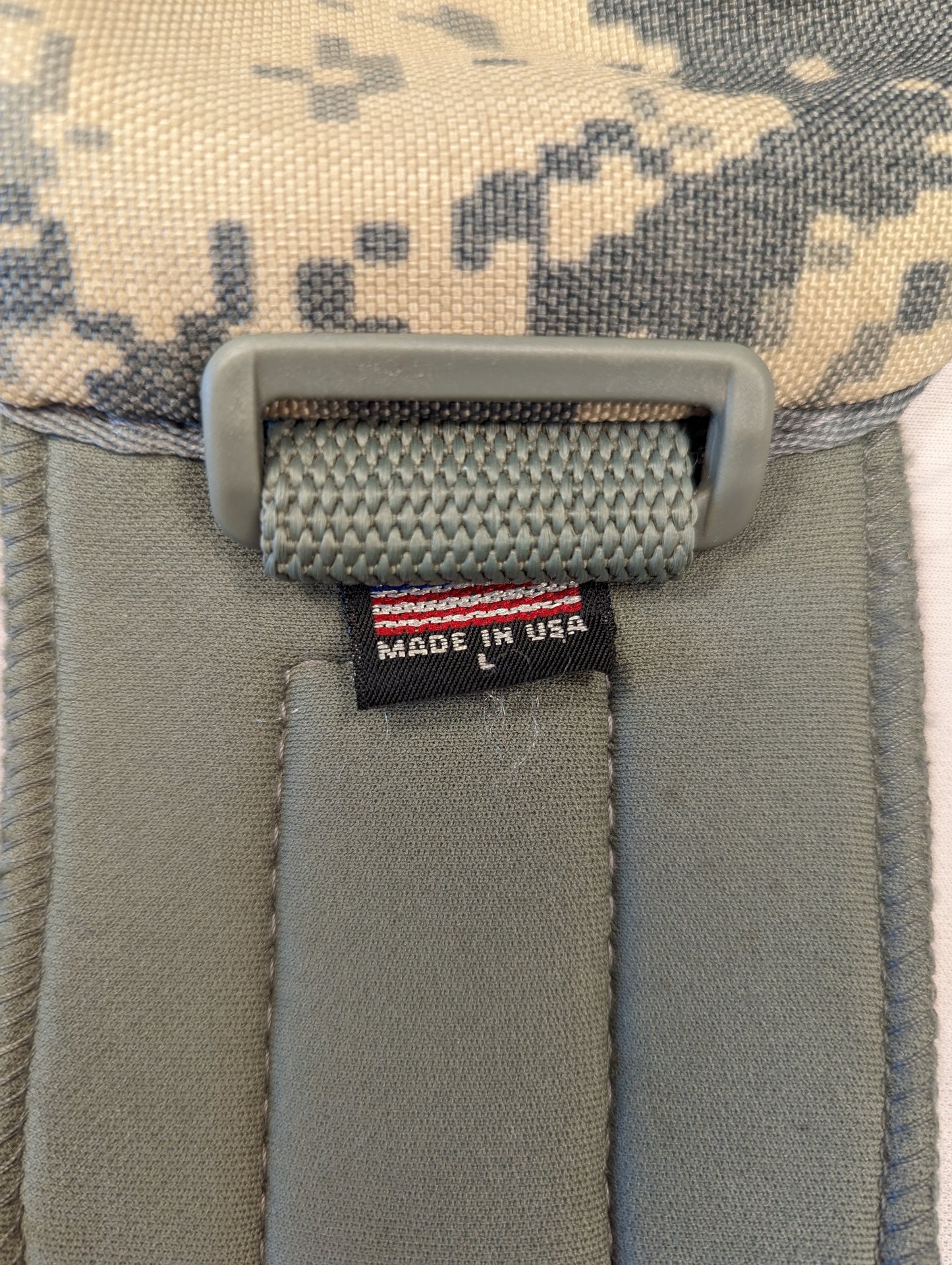 A close-up photograph of the 'MADE IN USA' tag with an American flag and size 'L' marking sewn onto the strap of a U.S. Military tactical pad, featuring ACU digital camouflage fabric.