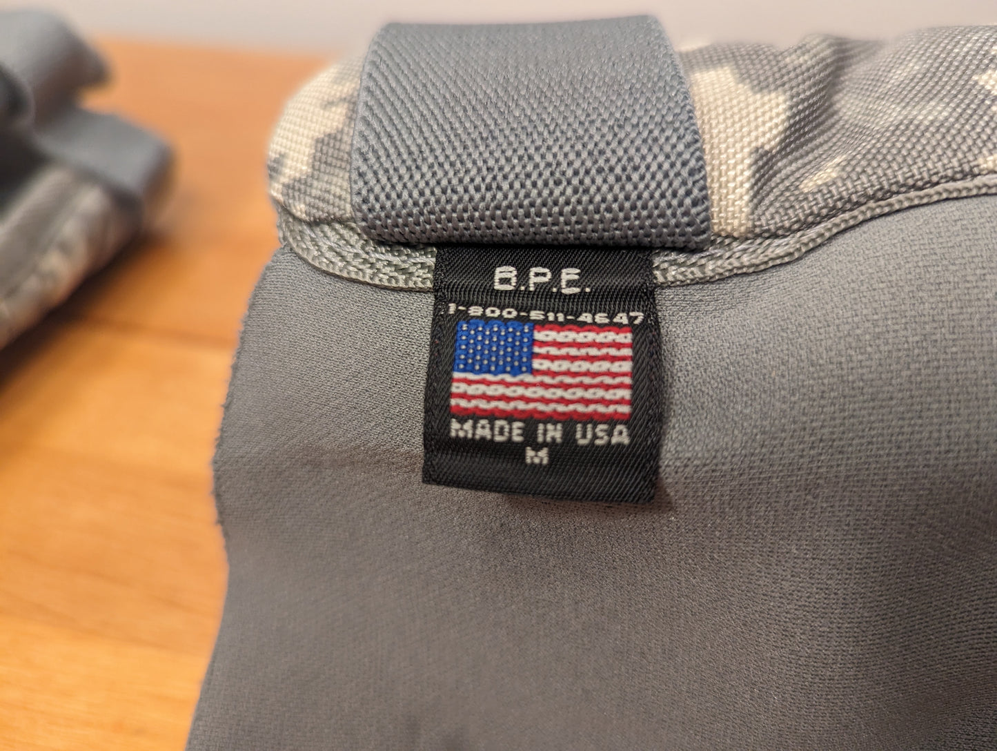 An extreme close-up photograph of the black 'MADE IN USA' tag sewn onto an ACU digital camouflage tactical pad strap, displaying the American flag graphic, manufacturer 'B.P.E.', and size 'M'.