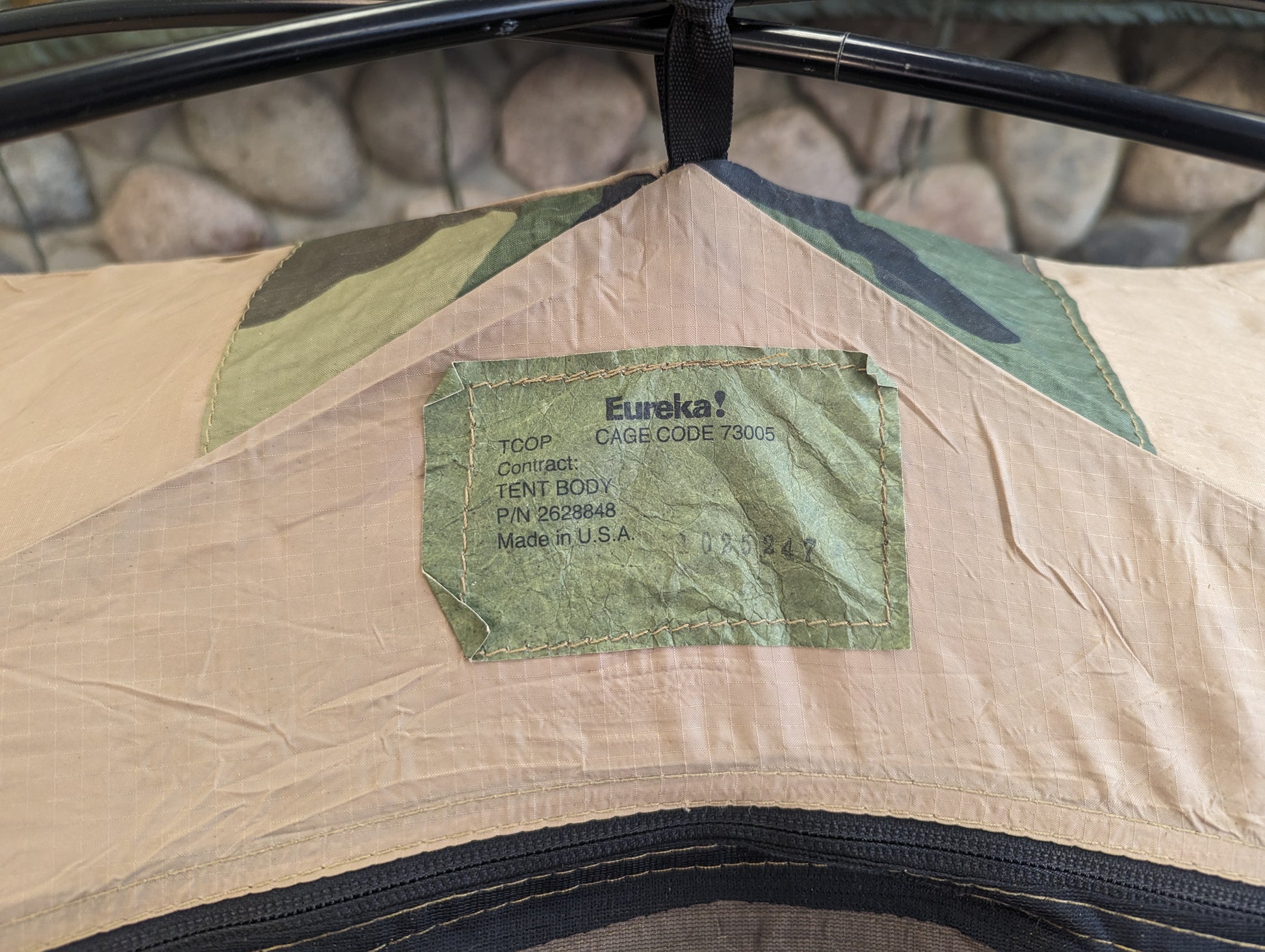 A close-up of the green manufacturer's tag sewn onto the tan ripstop fabric of the TCOP tent, displaying the 'Eureka!' logo, CAGE code, and 'Made in U.S.A.' text.