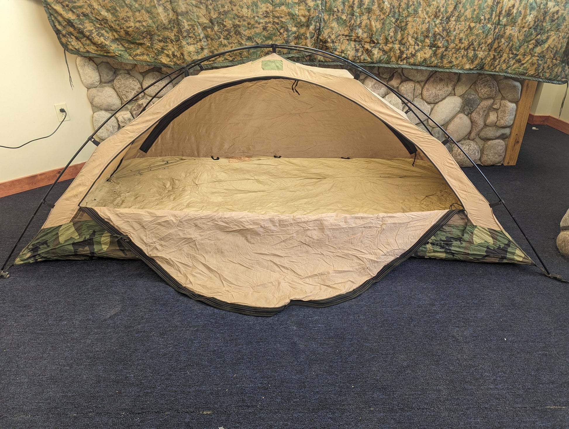 A front view of the tan inner body of the U.S. Military Combat Tent (TCOP) set up indoors, with the rainfly removed and the large door flap unzipped and laying flat to expose the interior sleeping area.