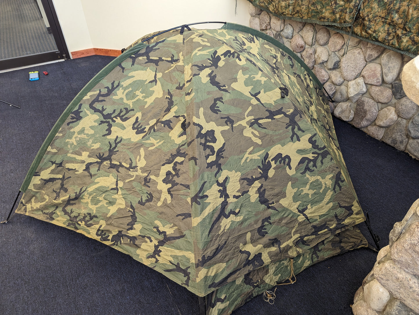 A single-person U.S. Military Surplus Combat Tent (TCOP) in Woodland camouflage, set up indoors on a carpeted floor, demonstrating its low-profile, compact design.