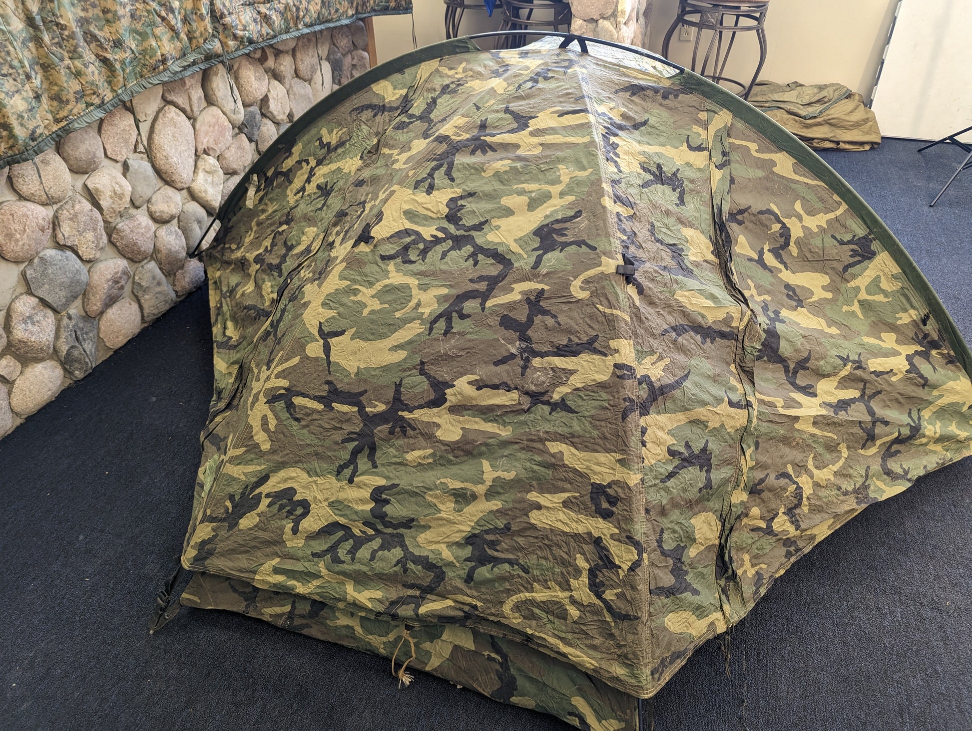 A single-person U.S. Military Surplus Combat Tent (TCOP) in Woodland camouflage, set up indoors on a carpeted floor, demonstrating its low-profile, compact design.