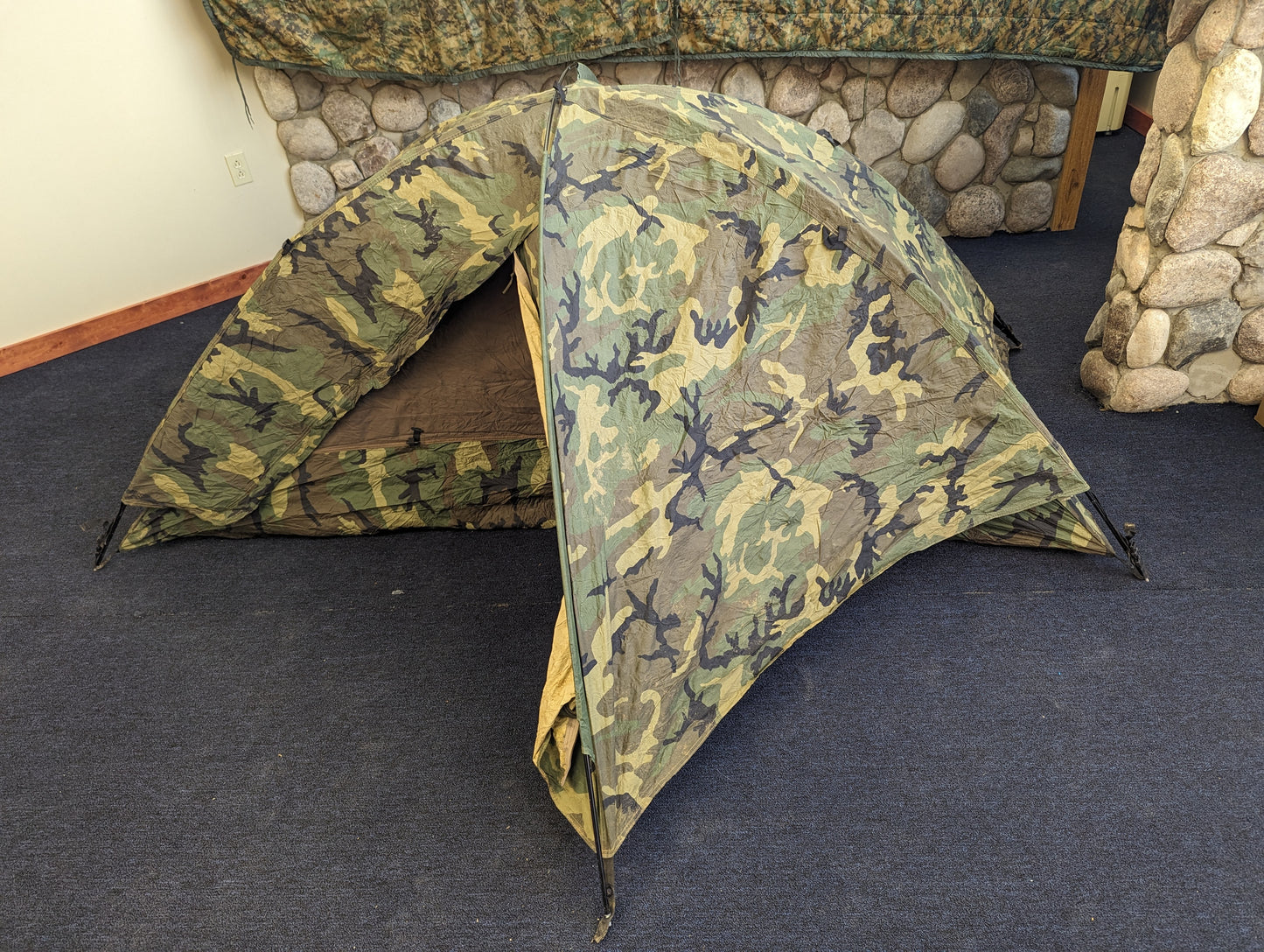 A single-person U.S. Military Surplus Combat Tent (TCOP) in Woodland camouflage, set up indoors on a carpeted floor, demonstrating its low-profile, compact design.