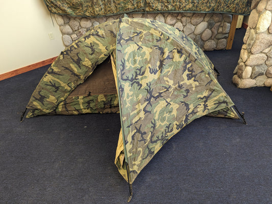A single-person U.S. Military Surplus Combat Tent (TCOP) in Woodland camouflage, set up indoors on a carpeted floor, demonstrating its low-profile, compact design.