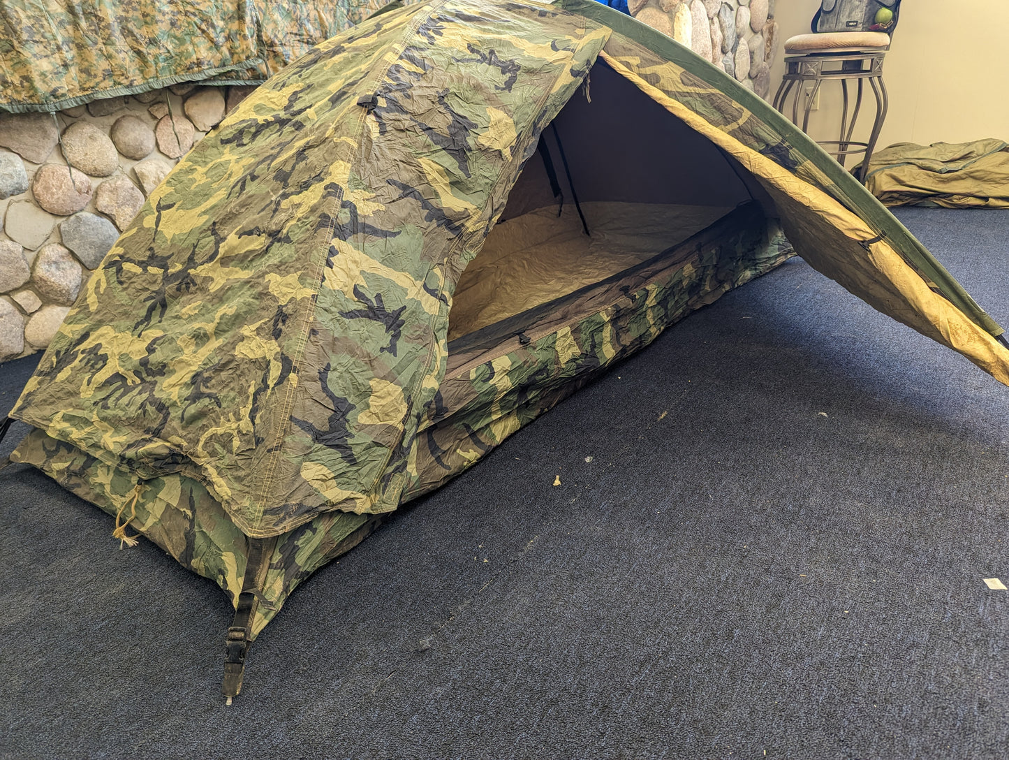 A single-person U.S. Military Surplus Combat Tent (TCOP) in Woodland camouflage, set up indoors with the door open to show the interior, resting on a dark carpeted floor.
