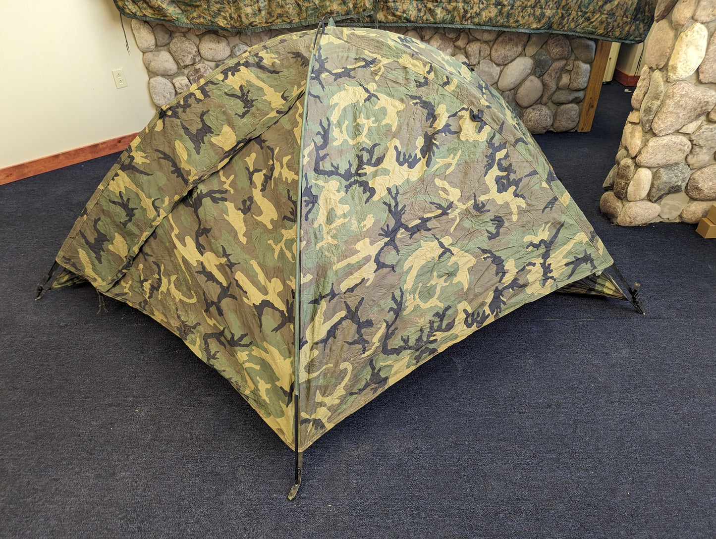 A single-person U.S. Military Surplus Combat Tent (TCOP) in Woodland camouflage, set up indoors on a carpeted floor against a stone wall, demonstrating its low-profile, compact design.