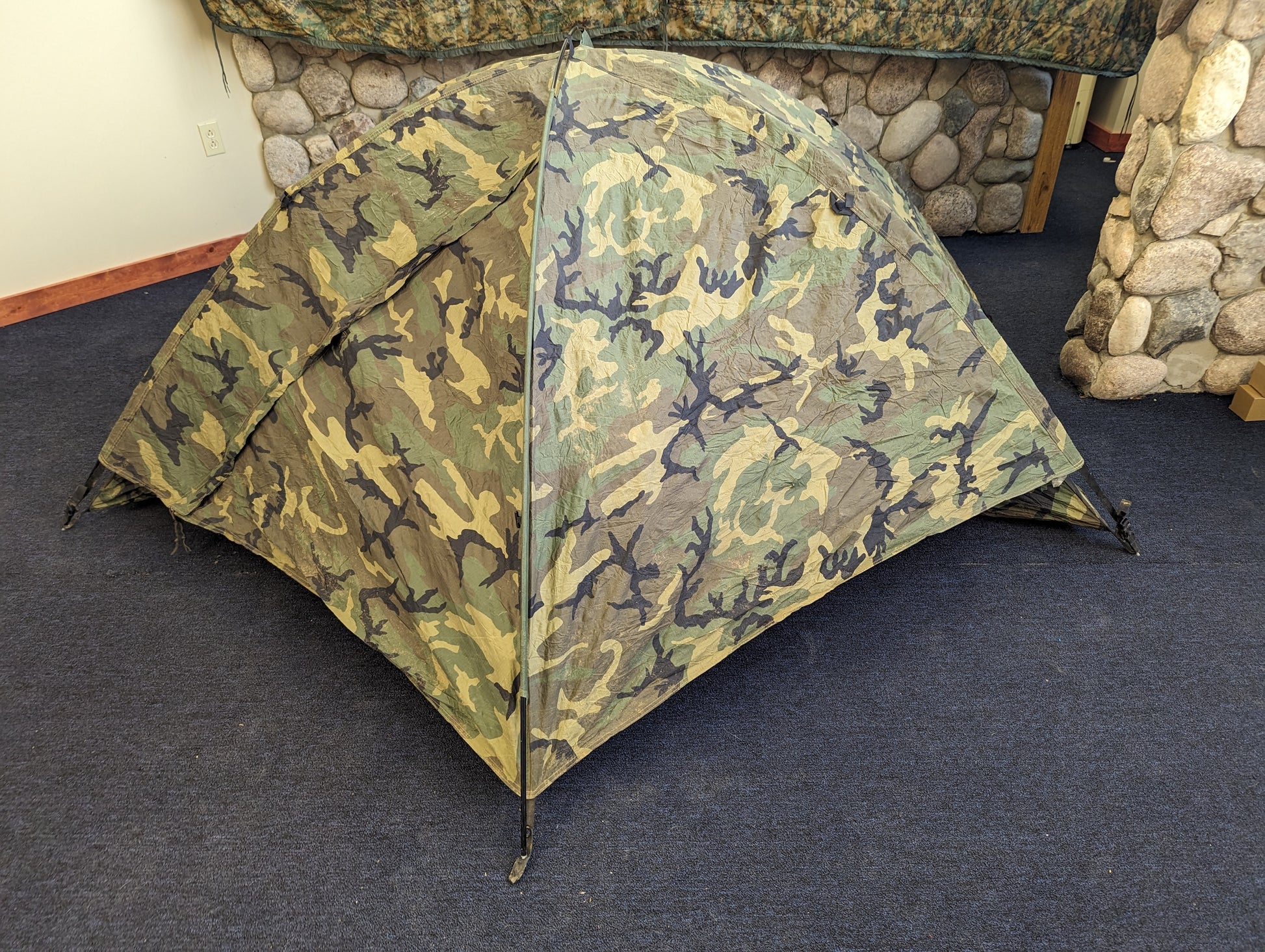 A single-person U.S. Military Surplus Combat Tent (TCOP) in Woodland camouflage, set up indoors on a carpeted floor against a stone wall, demonstrating its low-profile, compact design.