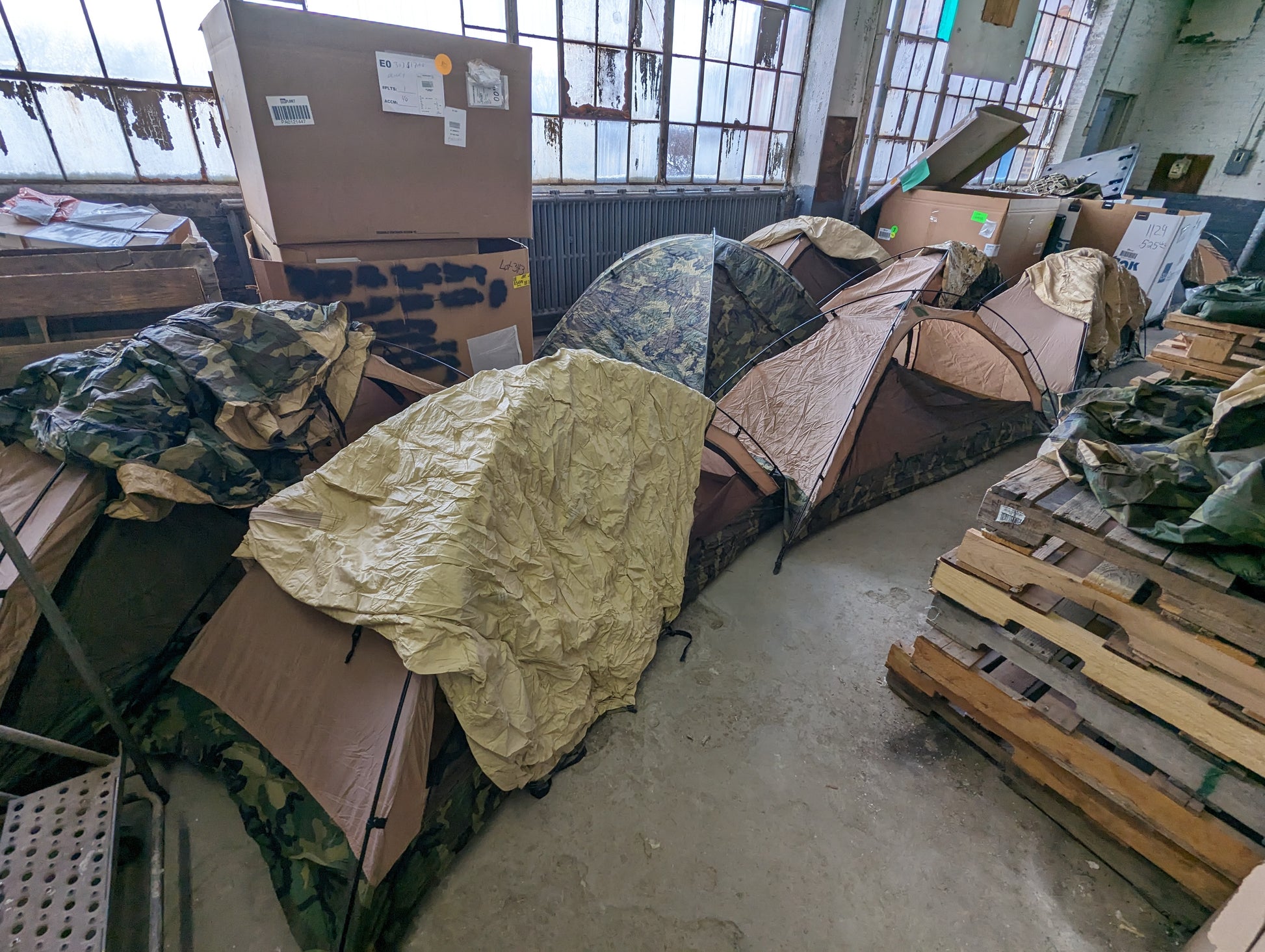 A high-angle view of a warehouse floor filled with multiple pitched U.S. Military Combat One-Person (TCOP) tents in Woodland camouflage and Tan, set up on wooden pallets amidst surplus boxes.
