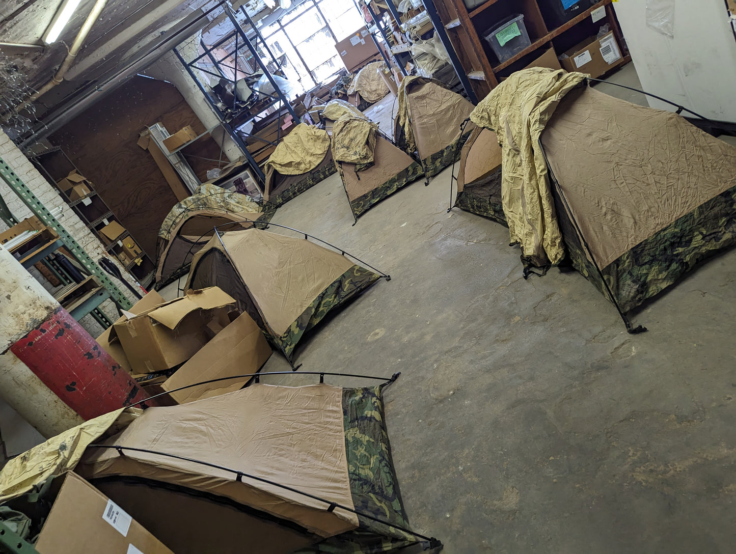 A high-angle view of a warehouse floor filled with multiple pitched U.S. Military Combat One-Person (TCOP) tents in Woodland camouflage, displaying the bulk surplus inventory.