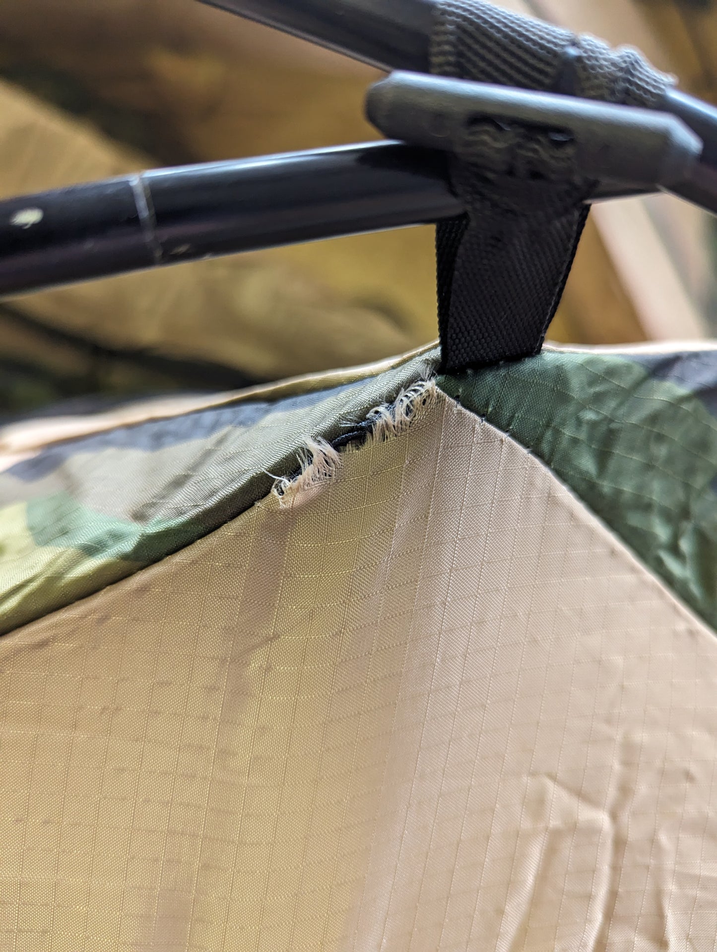 A close-up detail of the U.S. Military Combat Tent (TCOP) where the black metal frame connects to the Woodland camouflage fabric via a plastic fitting, showing wear and tear on the material.