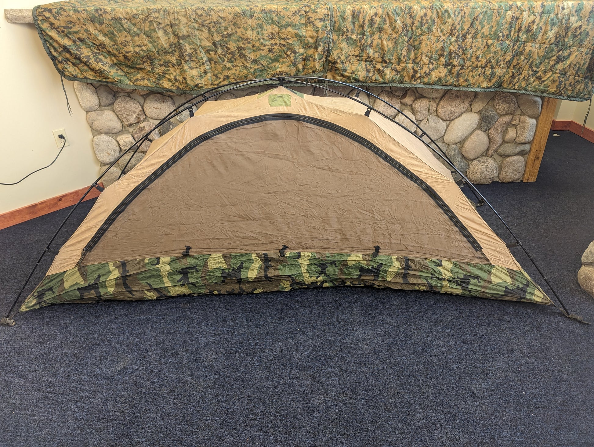 A close-up detail of the U.S. Military Combat Tent (TCOP) where the black metal frame connects to the Woodland camouflage fabric via a plastic fitting, showing the seams and material condition.