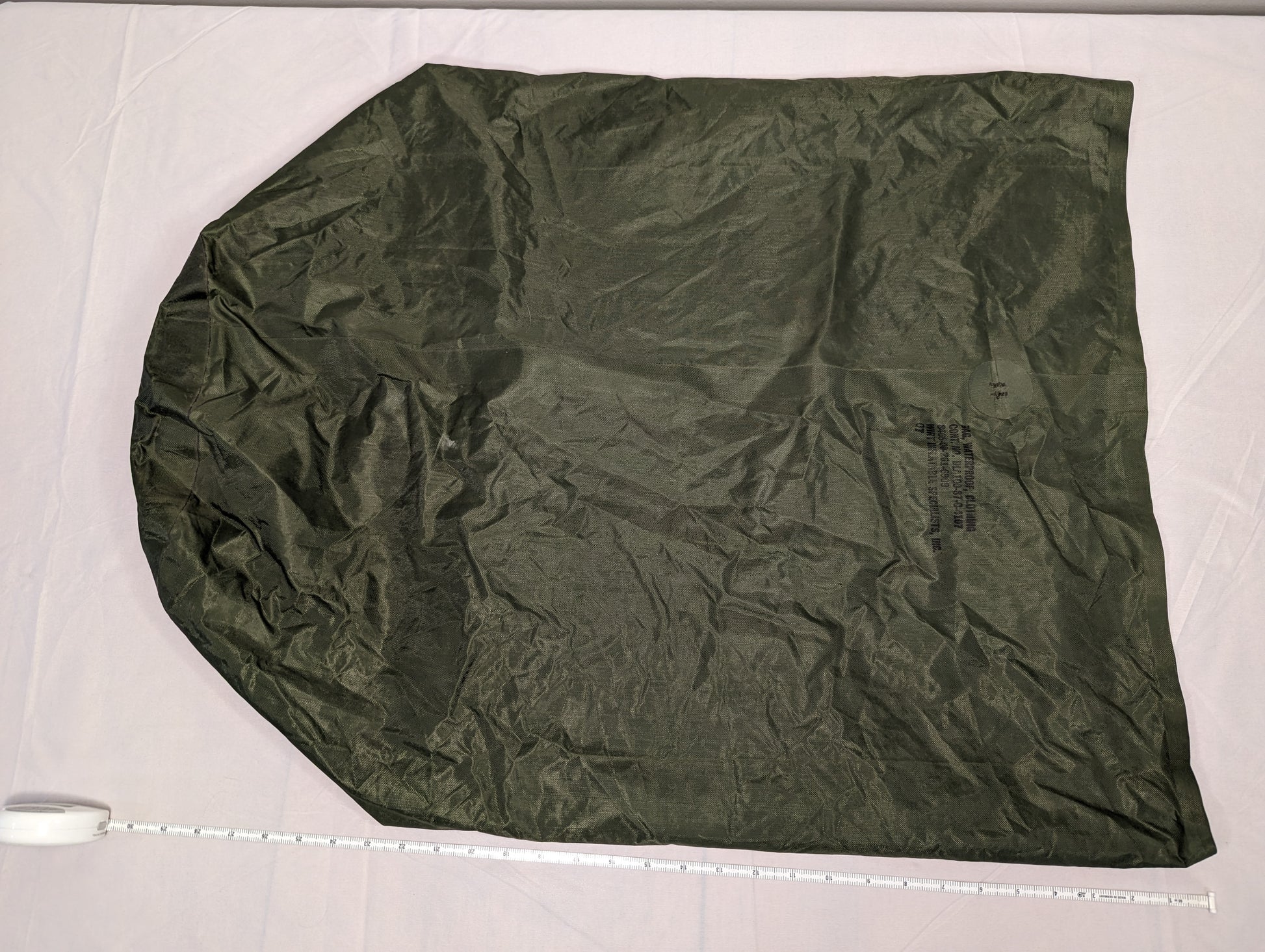 A large, fully unfolded U.S. Military Waterproof Clothing Bag in olive green ripstop nylon, laid flat on a white surface with a tape measure placed at the base to indicate the bag's dimensions.