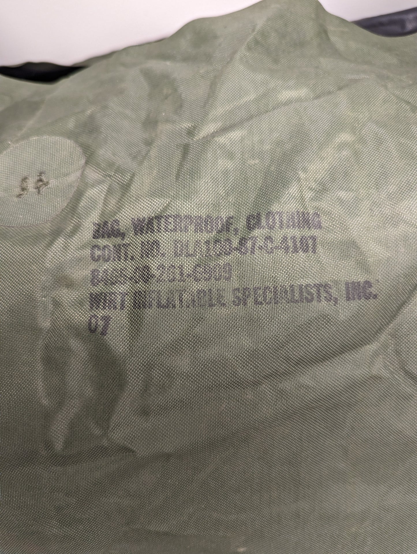 A close-up photograph of a U.S. Military Waterproof Clothing Bag in olive green, displaying the black stamped identification text, including 'BAG, WATERPROOF, CLOTHING', the NSN, contract number, and manufacturer details.