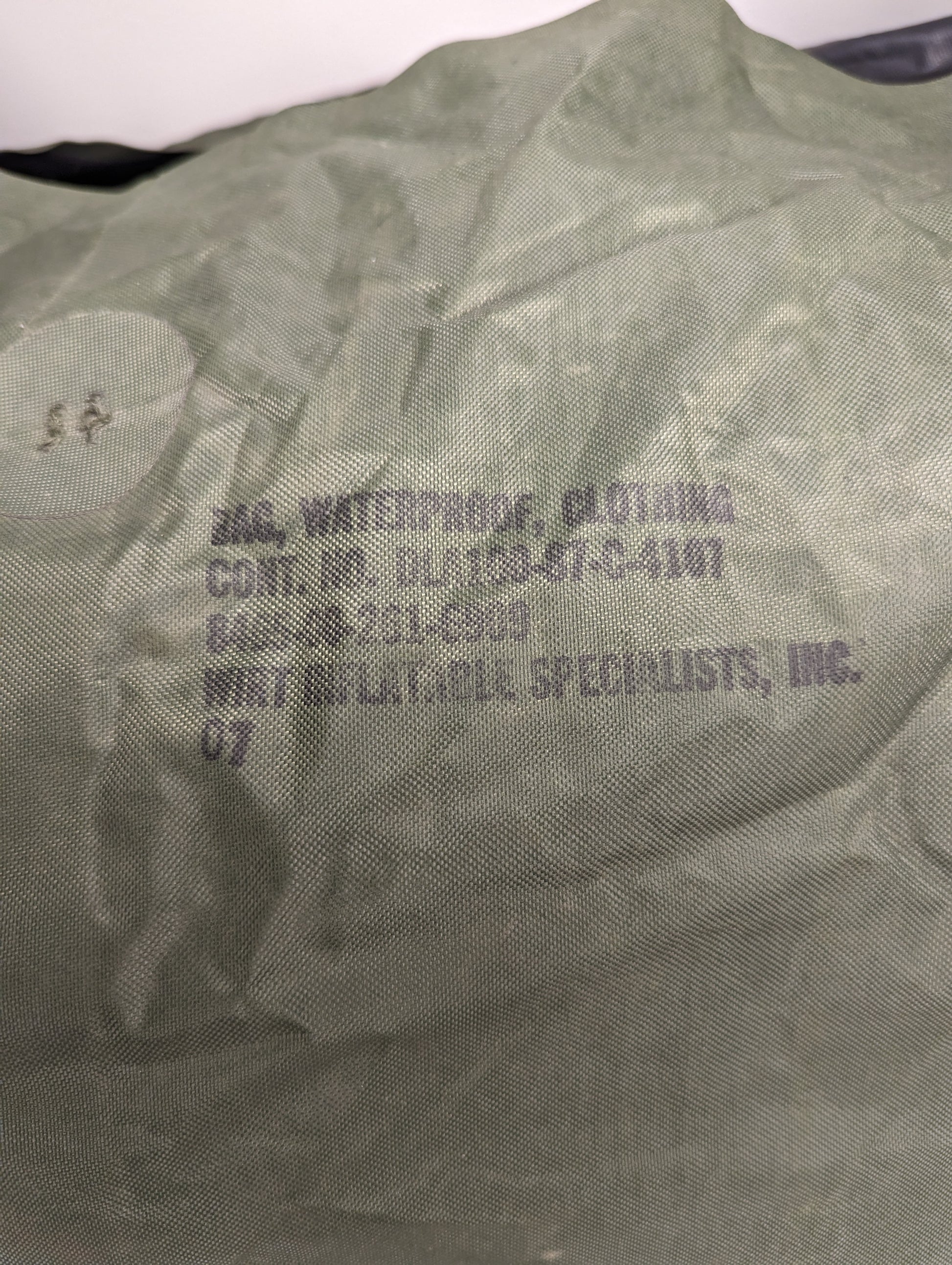A close-up photograph of a U.S. Military Waterproof Clothing Bag in olive green, displaying the black stamped identification text, including 'BAG, WATERPROOF, CLOTHING', the NSN, contract number, and manufacturer details.