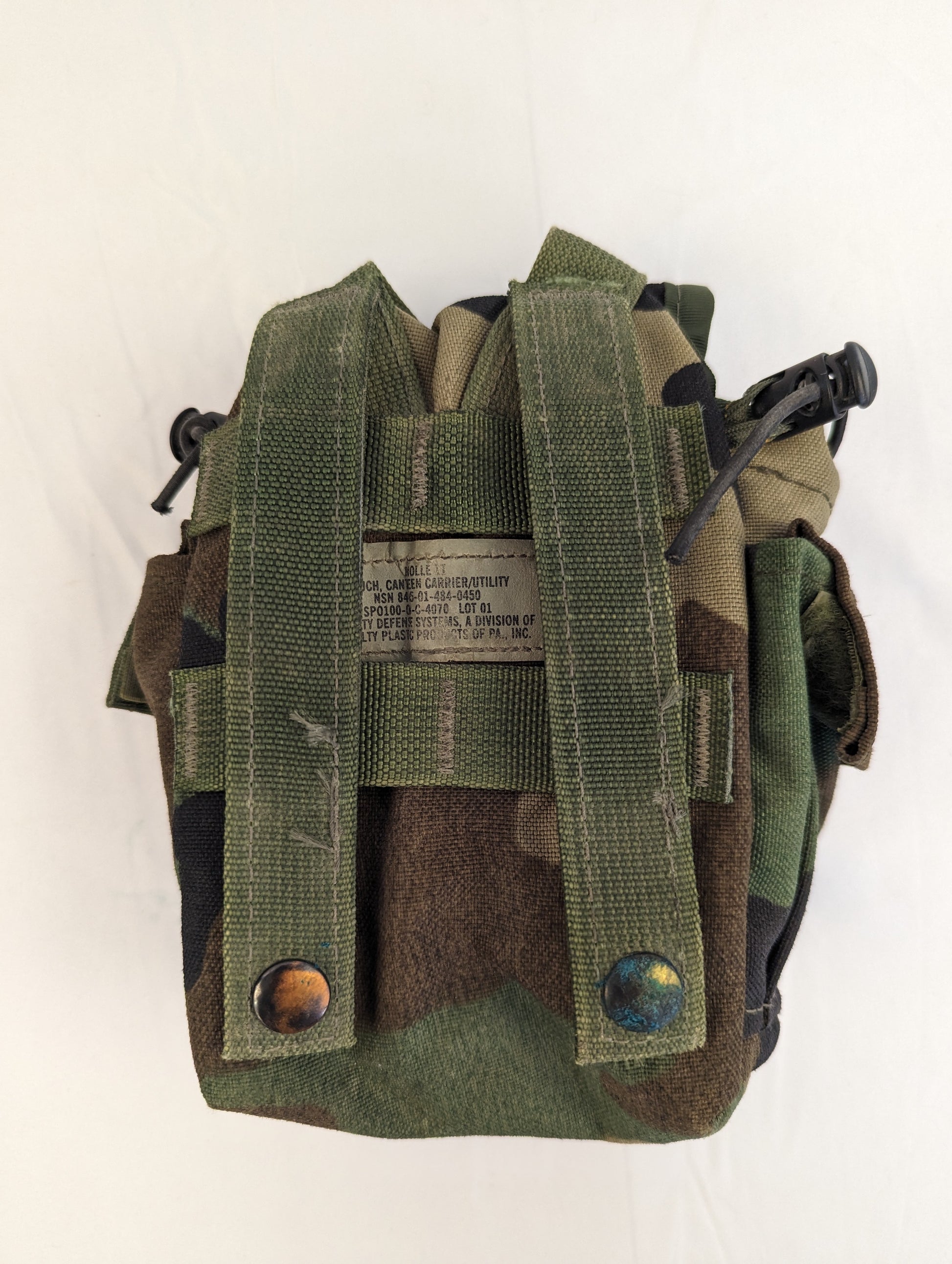 A close-up photograph of the back of a U.S. Military Surplus 1-Quart Canteen Pouch in Woodland camouflage, displaying the vertical ALICE/MOLLE attachment straps and a stitched manufacturer's label.