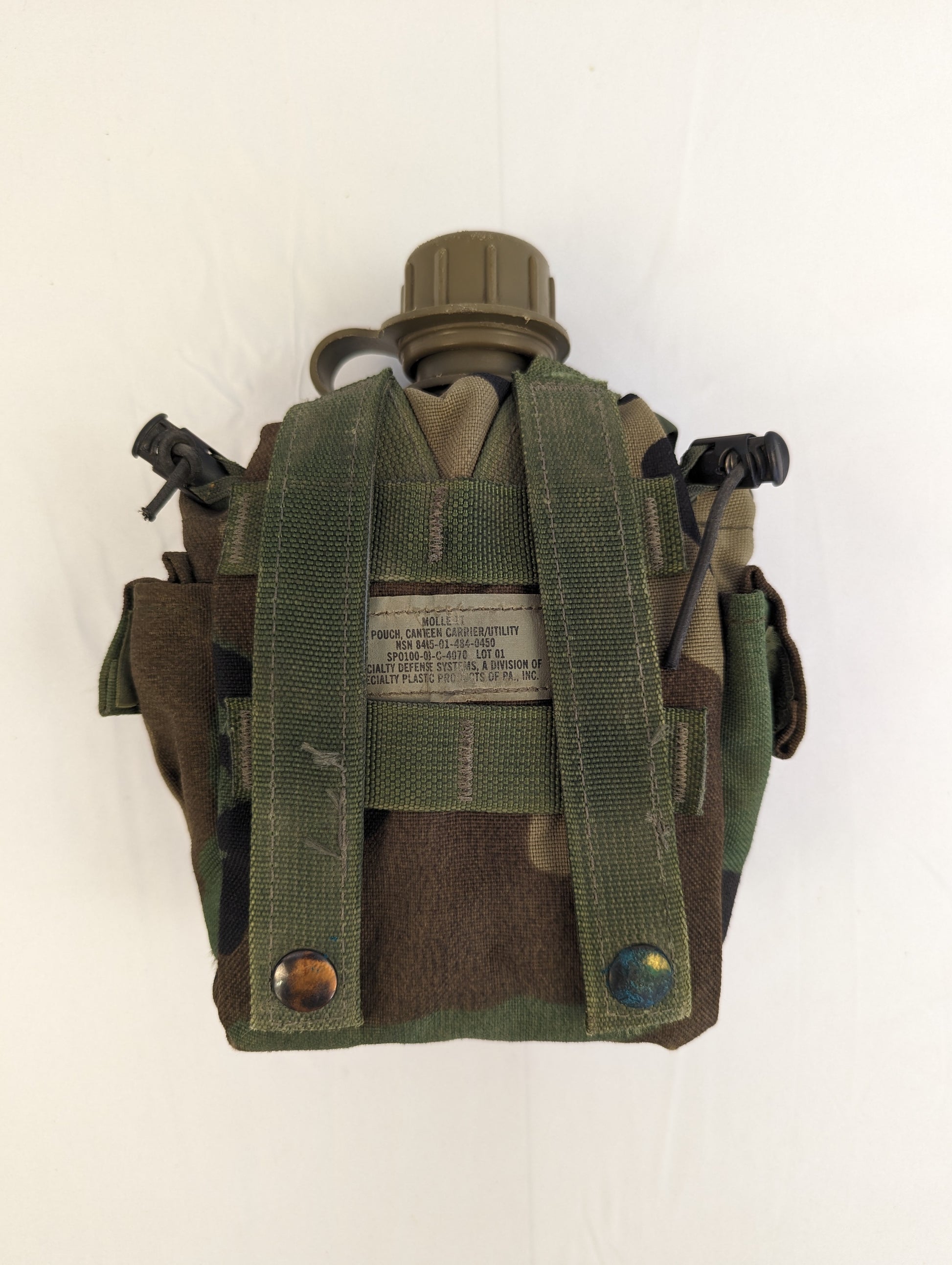 A back view of a U.S. Military Surplus Woodland camouflage 1-quart canteen pouch, displaying the vertical MOLLE attachment webbing, two drainage grommets, and the stitched manufacturer's label.