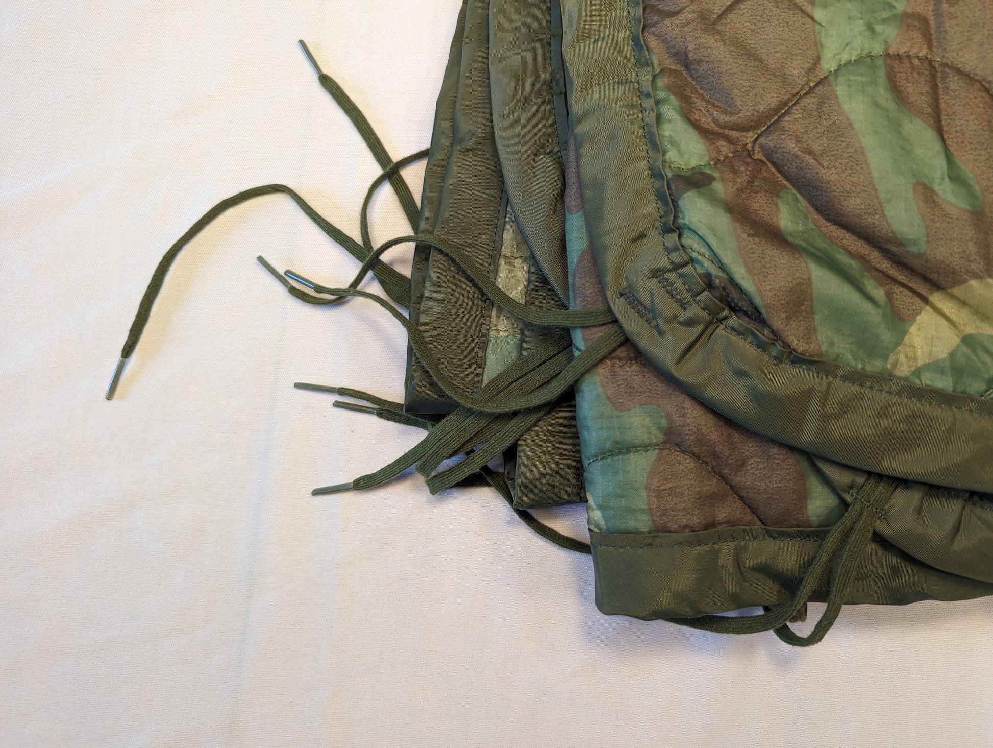 An extreme close-up detail shot of the corner of a U.S. Military Woodland Camouflage Poncho Liner, highlighting the quilted pattern, dark green edge binding, and several loose attachment tie cords.