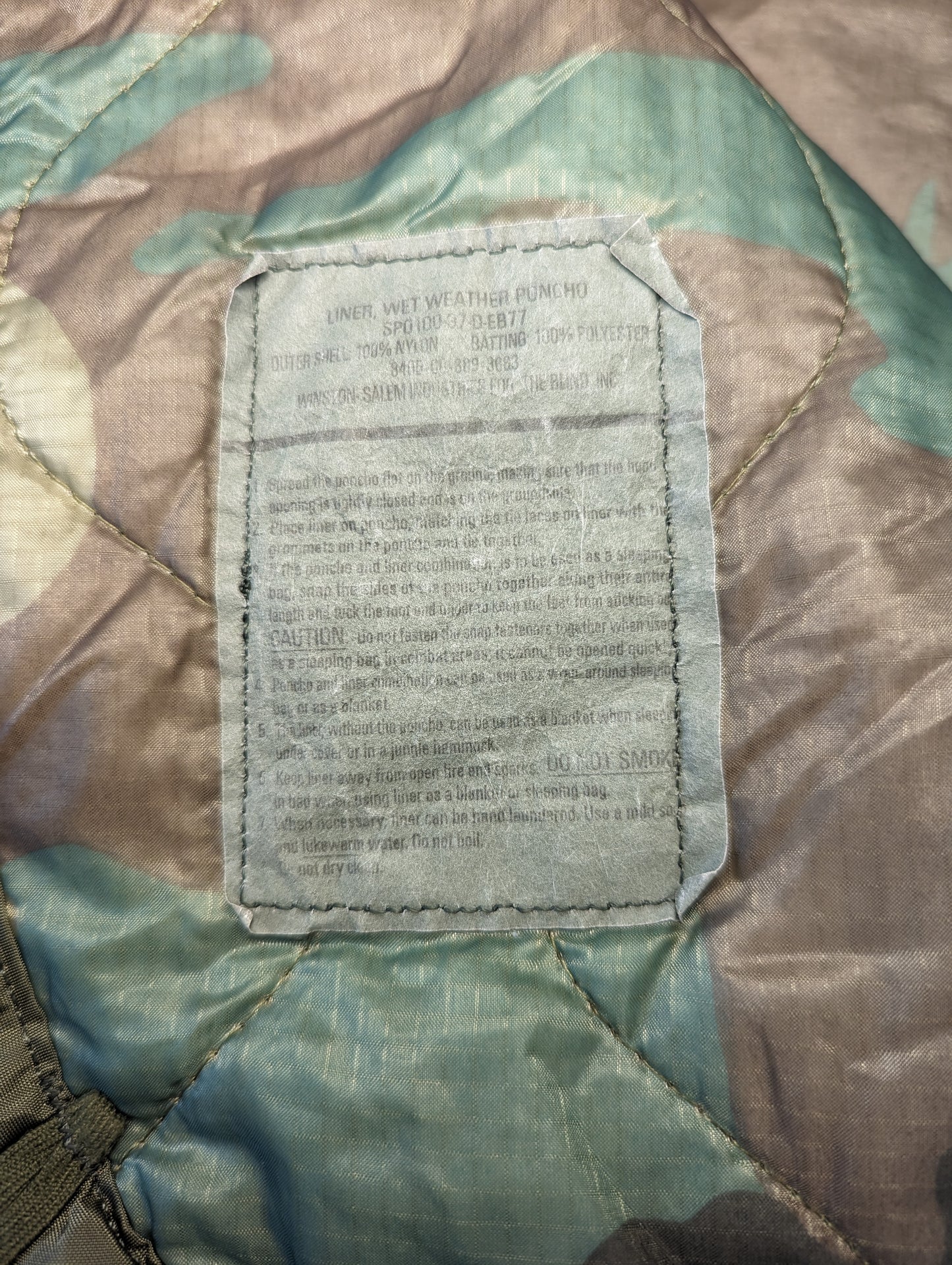 A close-up photograph of the large white military issue and care label sewn onto the Woodland camouflage Poncho Liner, displaying dense blocks of faded text and detailed laundering instructions.