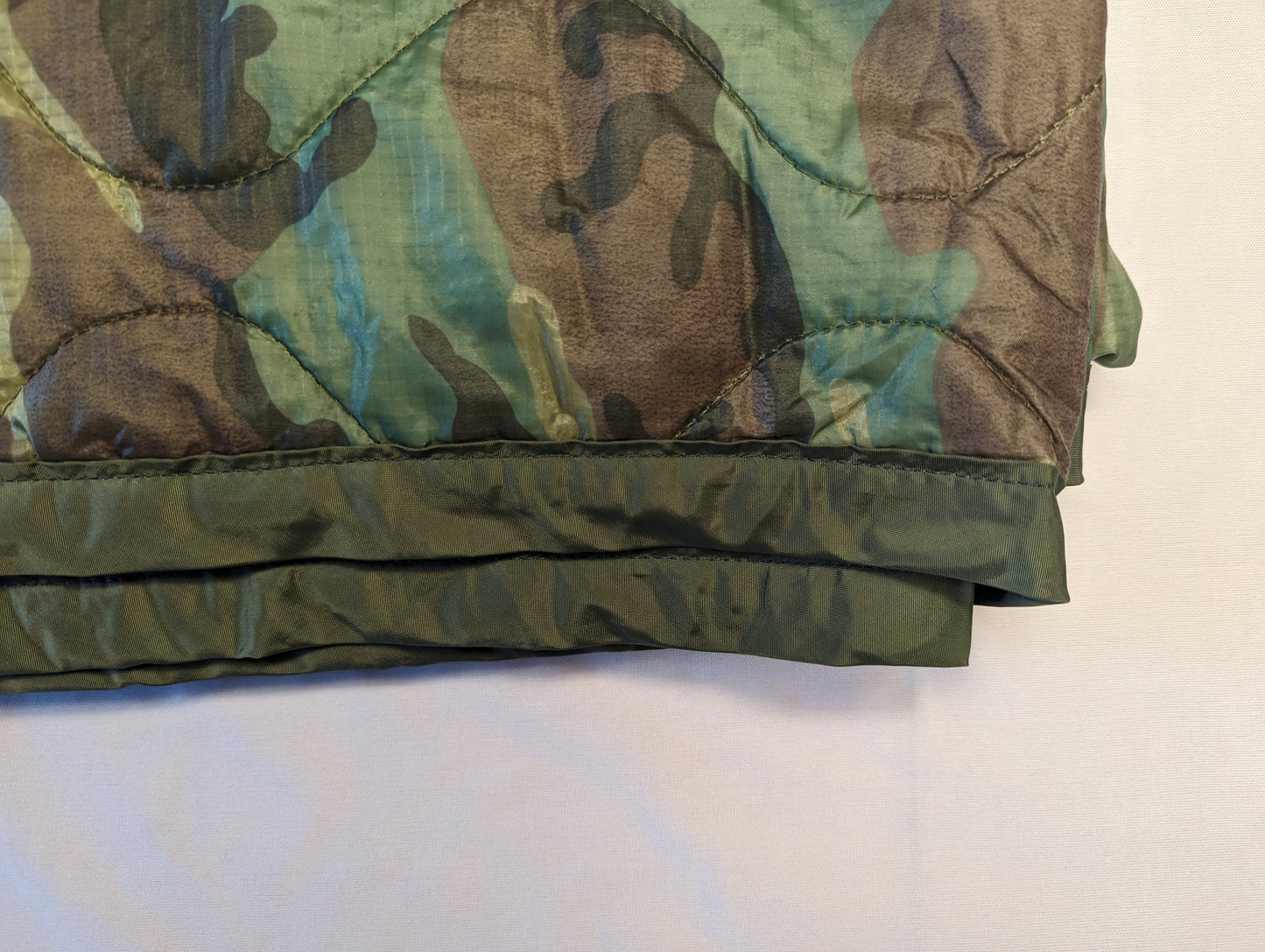 A close-up view of the bottom edge of a U.S. Military Woodland camouflage Poncho Liner (Woobie), highlighting the quilted fabric texture and the heavy, dark green binding.